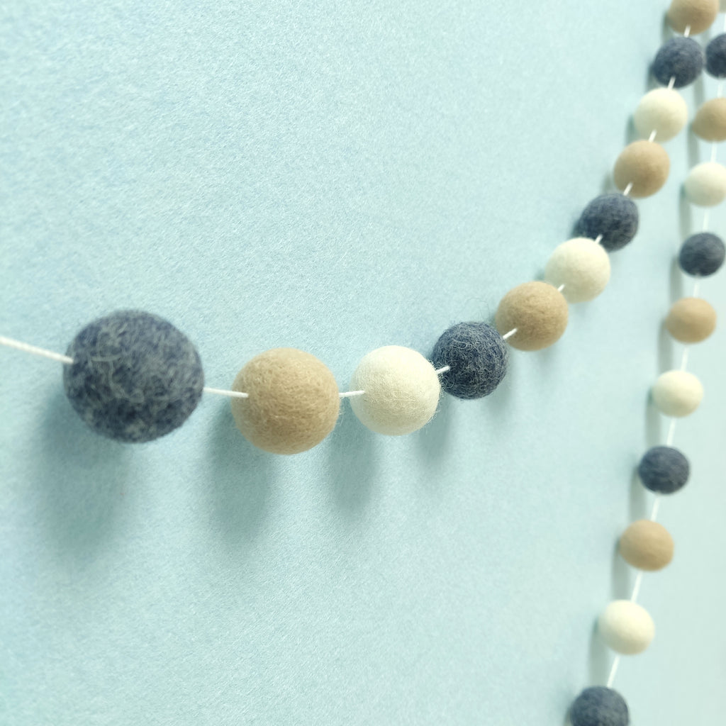 Guirlande de pompons en feutre denim beige et blanc vue de profil sur un mur