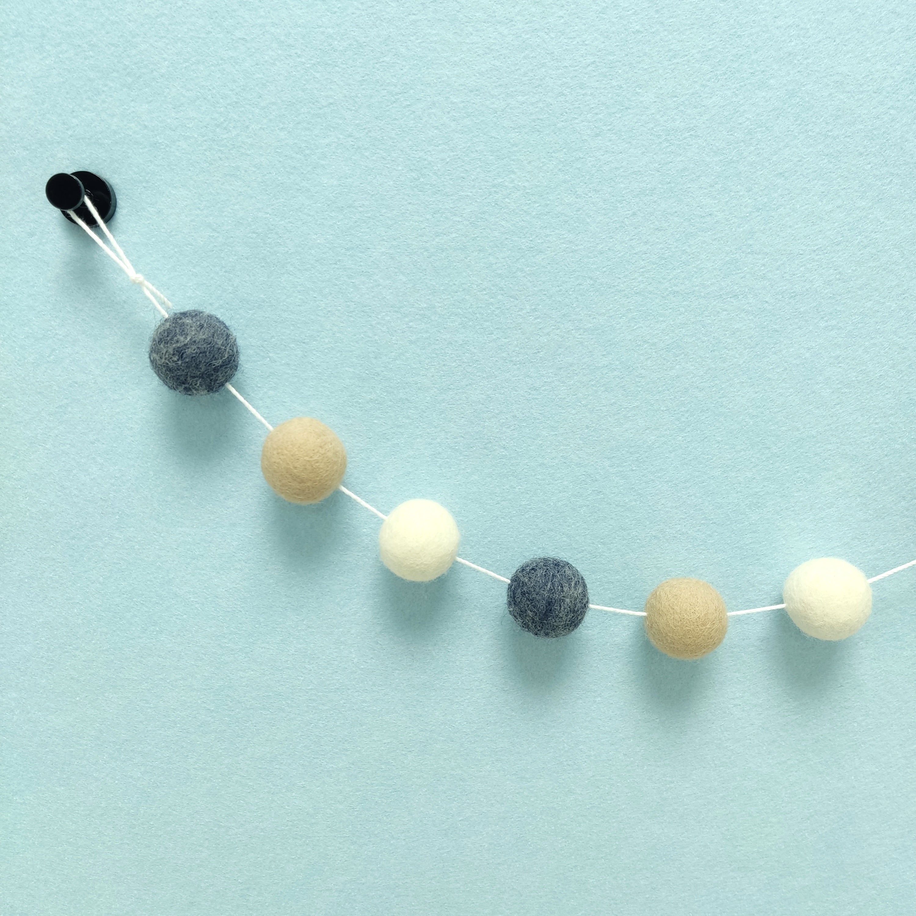 Zoom sur les couleurs de la guirlande de pompons bleu jeans brut, beige poudré et blanc naturel avec la boucle de fixation
