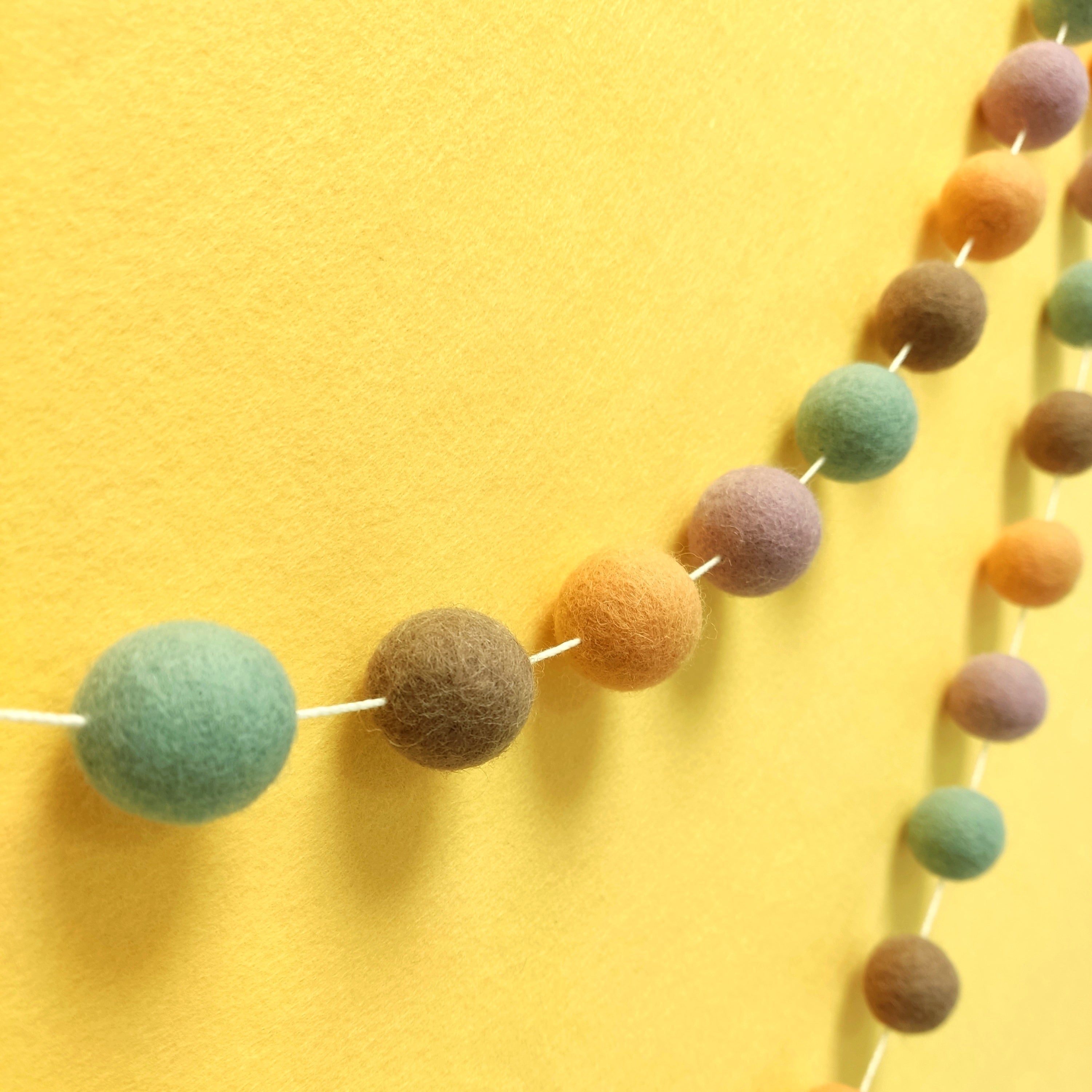 Guirlande de pompons en feutre marron vert lilas orange vue de profil sur un mur