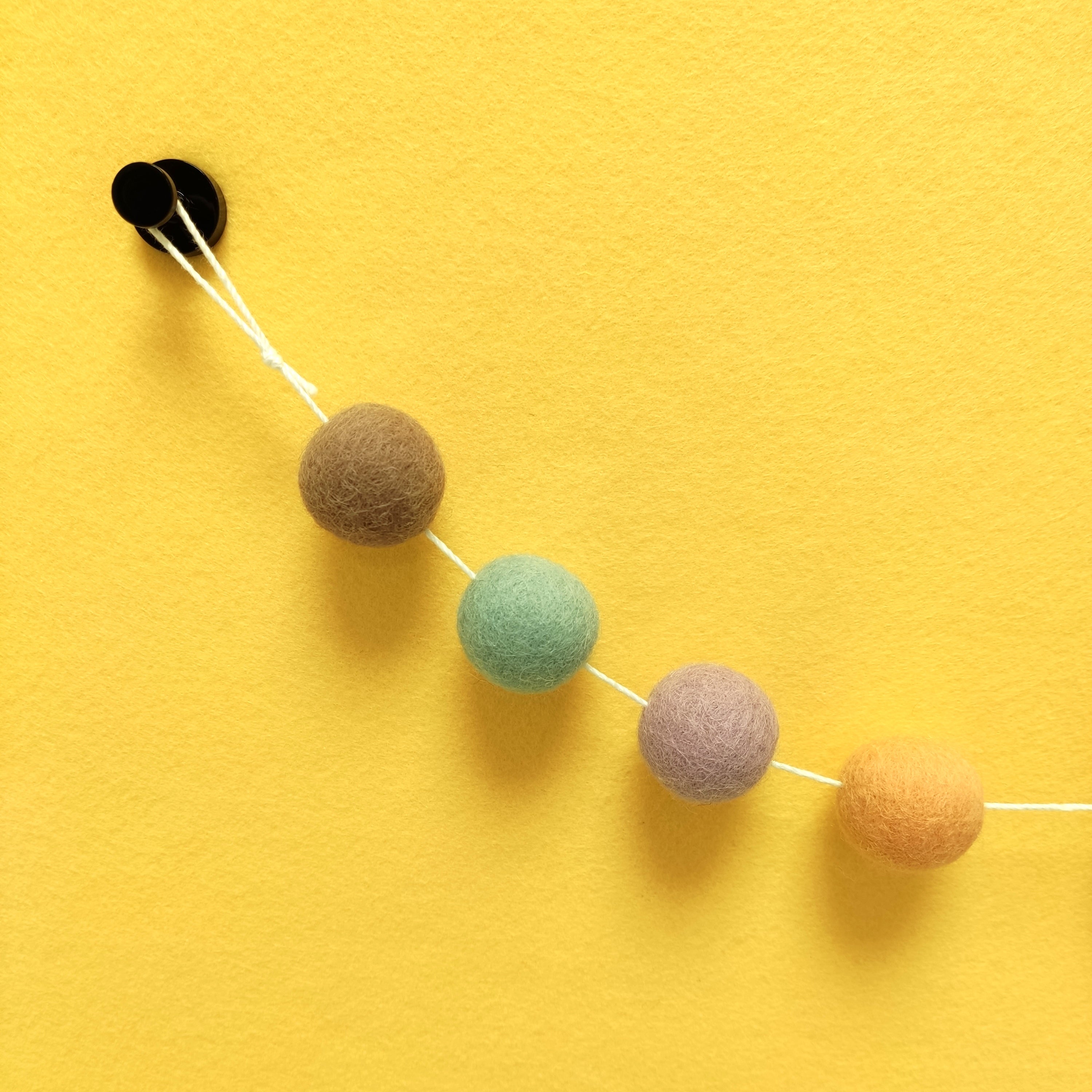 Zoom sur les pompons feutrés beige cappuccino, lilas, orange pêche et vert céladon de la guirlande dolce vita