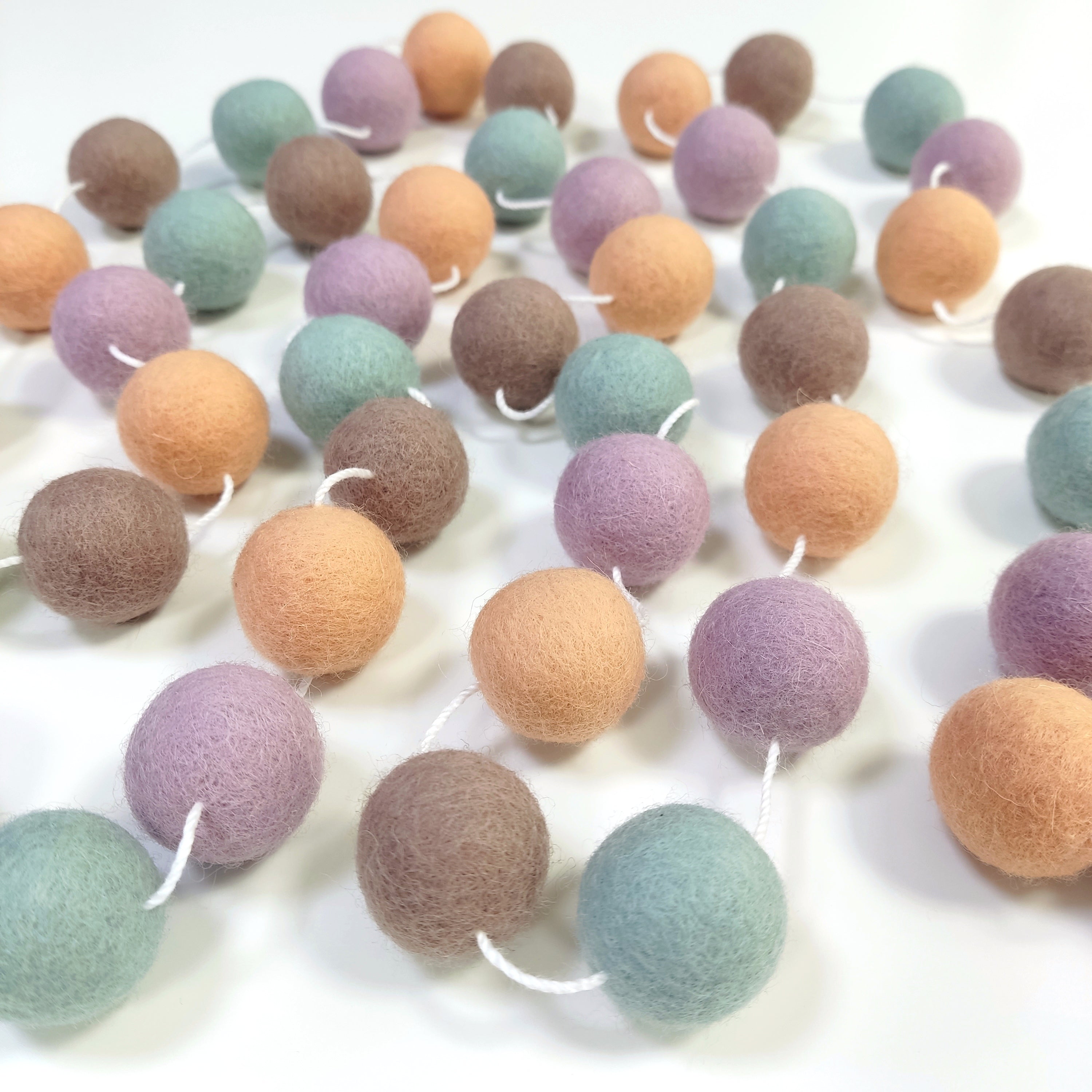 Guirlande mauve lilas, vert céladon, beige cappuccino et orange pêche, étalée sur une table avec les pompons espacés