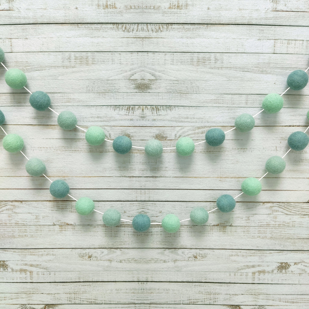 Guirlande de pompons feutrés aux teintes vert bleu eucalyptus suspendue sur un mur