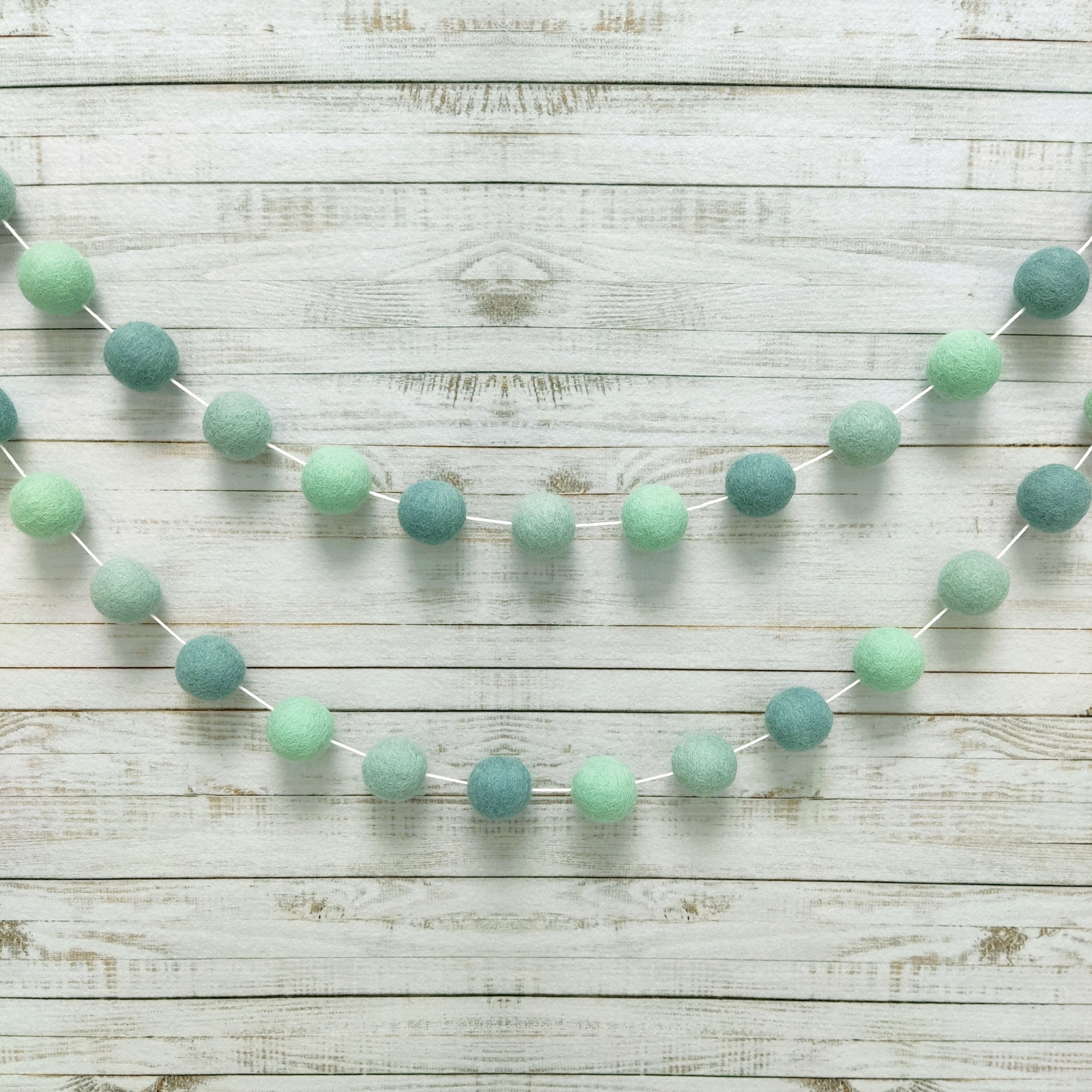 Guirlande de pompons feutrés aux teintes vert bleu eucalyptus suspendue sur un mur