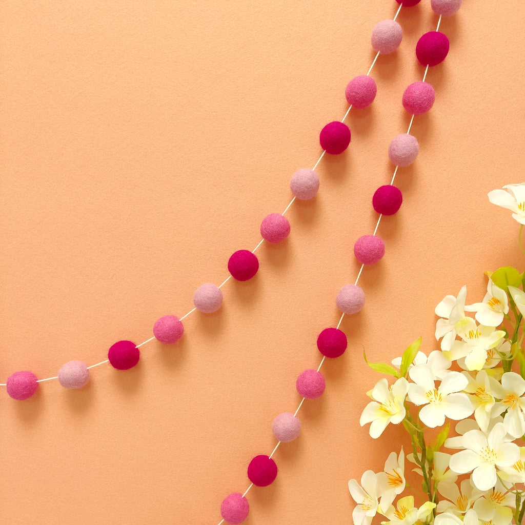 Guirlande de pompons feutrés en laine mérinos rose magenta, rose bonbon et rose cerisier accroché sur un mur