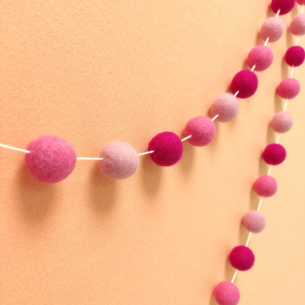 Guirlande de pompons en laine feutrée rose bonbon, rose cerisier et rose magenta vue de profil sur un mur