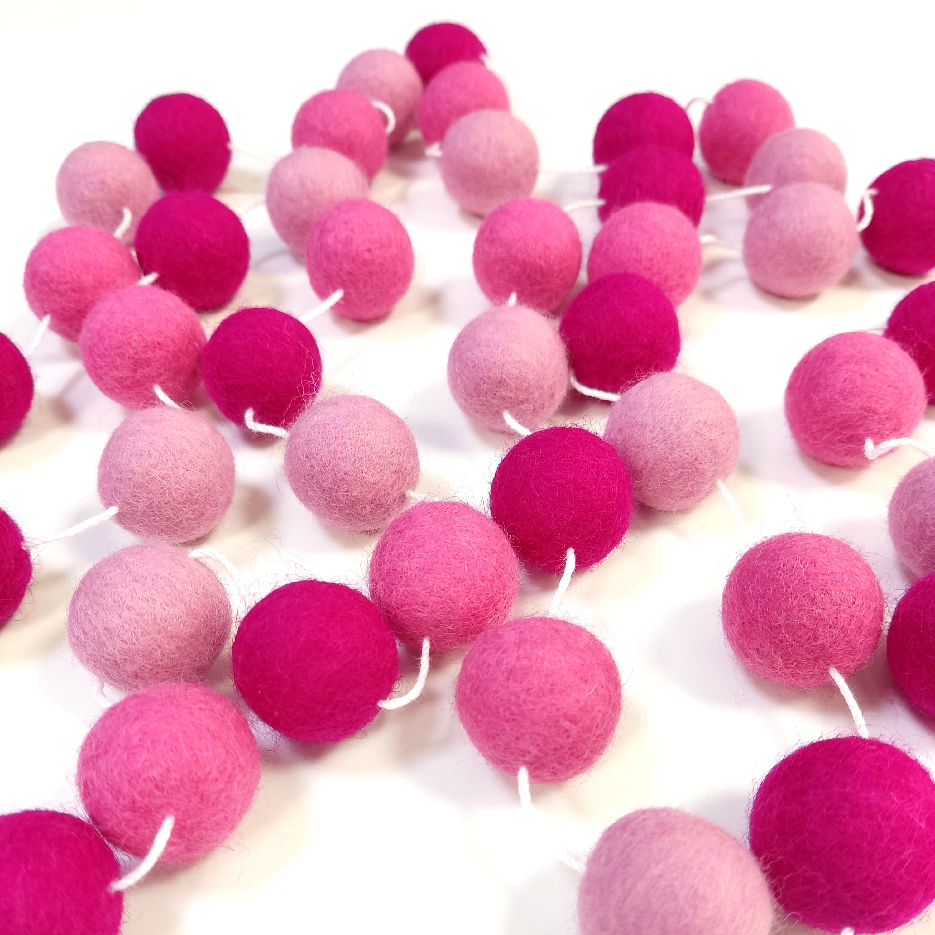 Guirlande en boules de laine feutrée rose magenta, rose bonbon et rose cerisier étalée sur une table avec les pompons espacés
