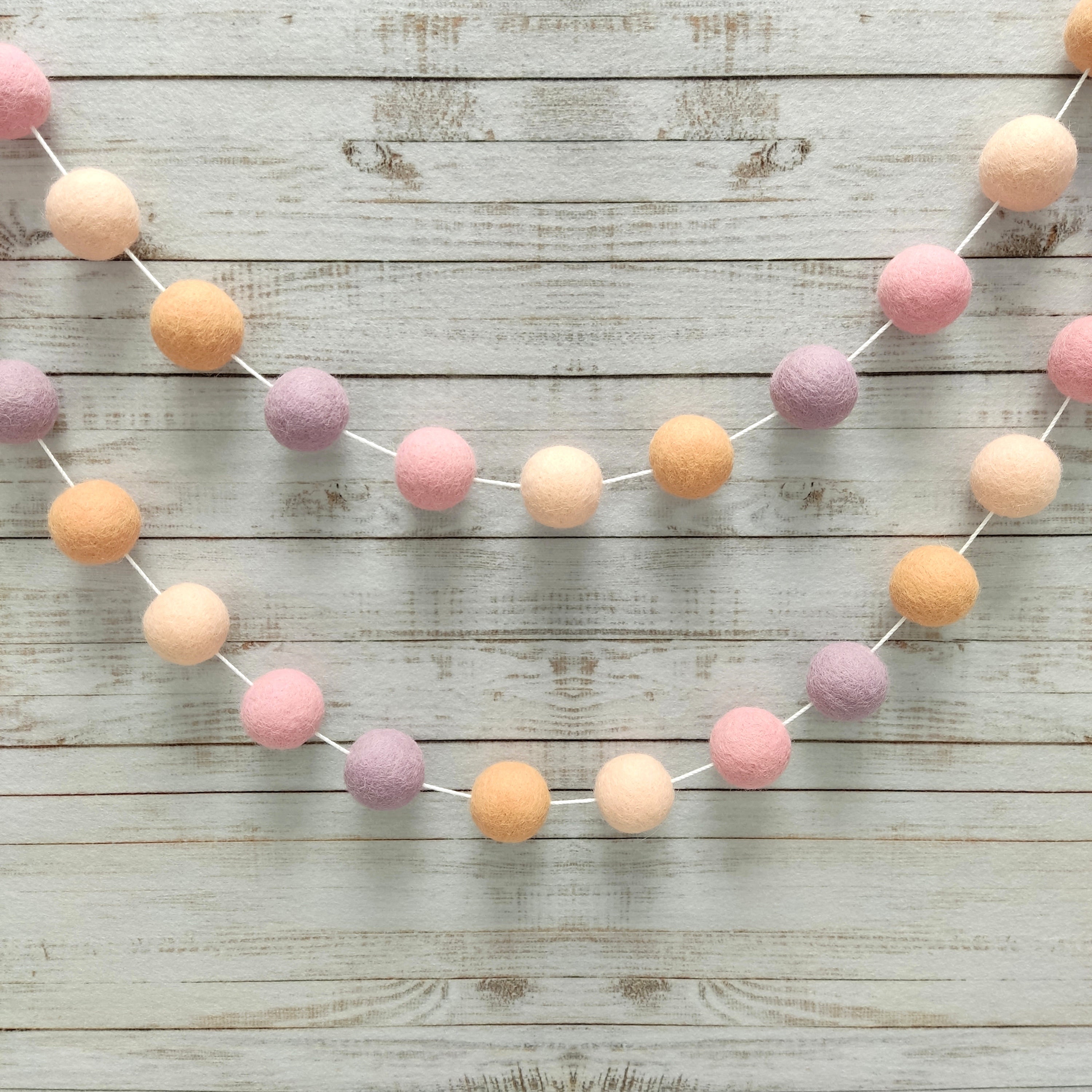 Guirlande pompons en feutre girly pêche, rose et lilas fixée sur un mur vue de près