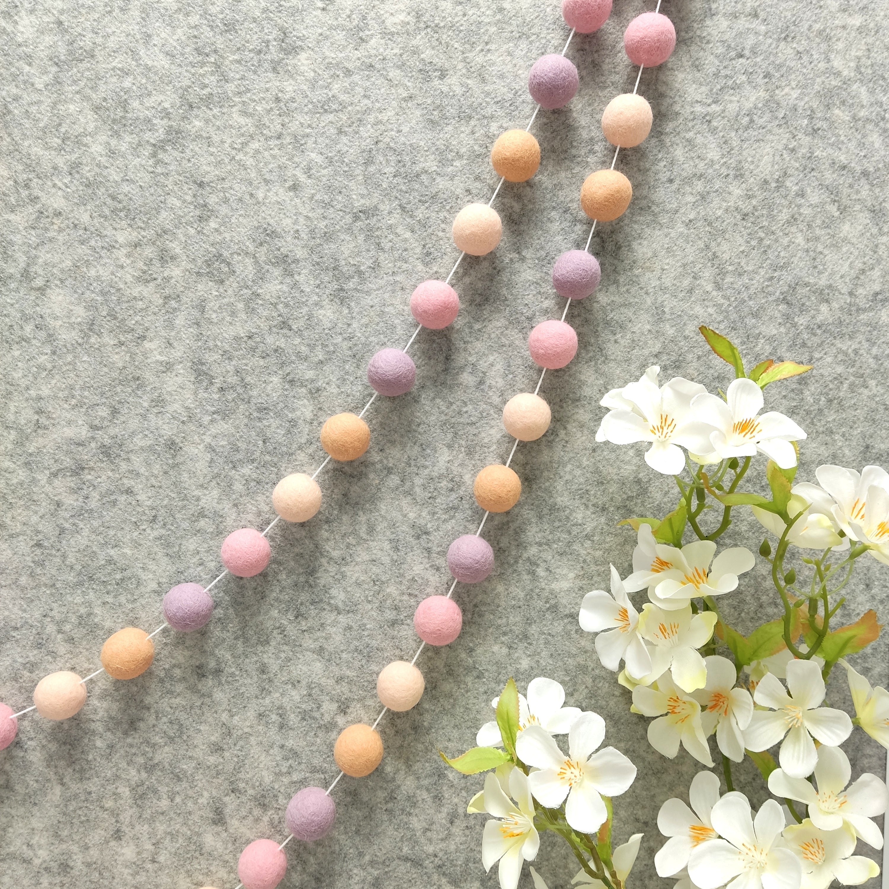 Guirlande de pompons rose, lilas et orange pêche suspendue sur un mur
