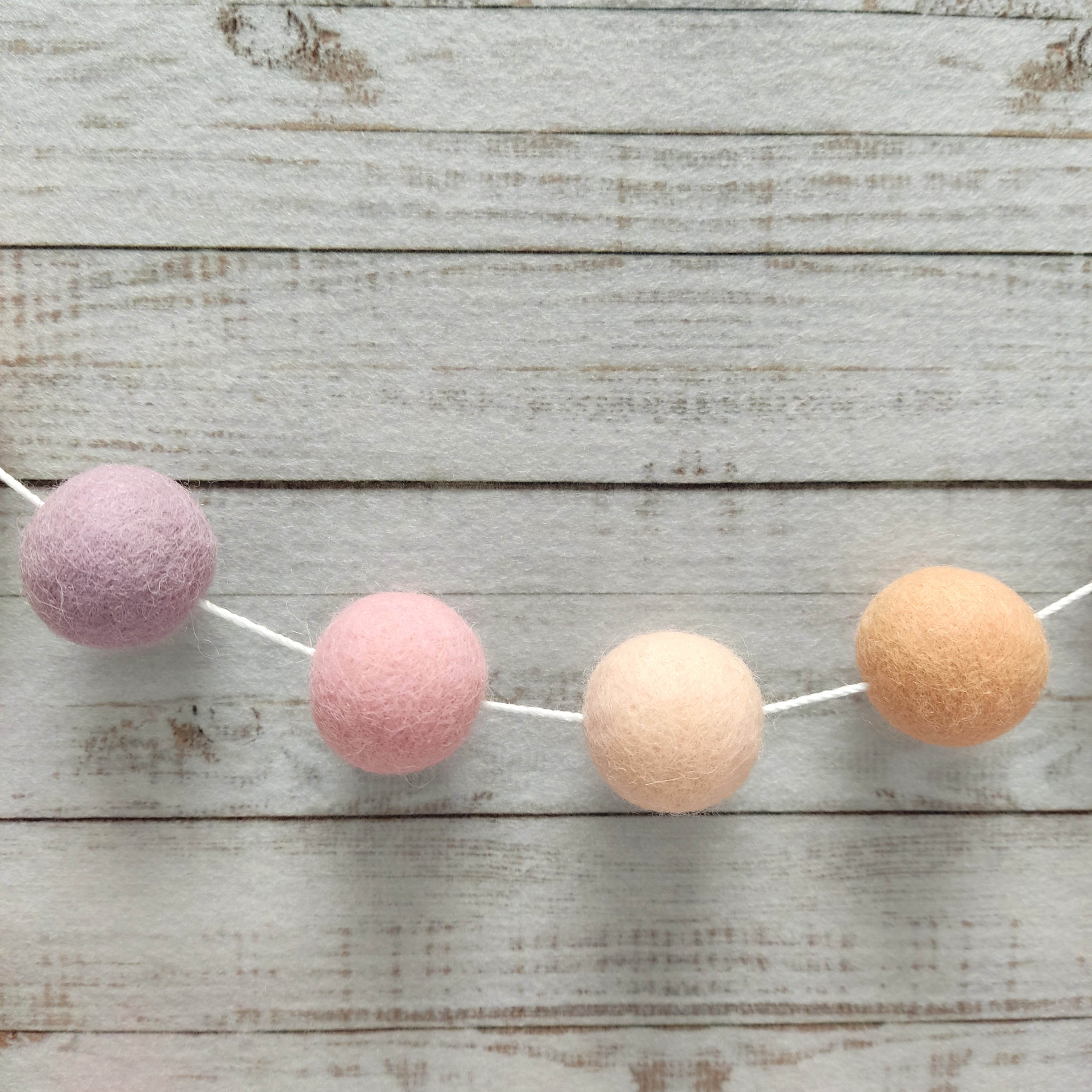 Zoom sur les pompons rose cerisier, orange pêche, beige rosé et lilas de la guirlande girly pêche en boules de feutre