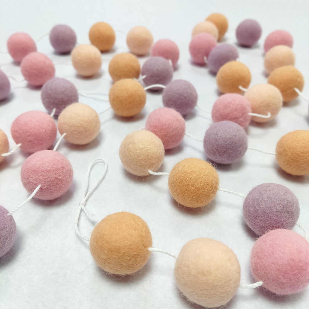 Guirlande de pompons en feutre rose lilas et orange posée sur une table