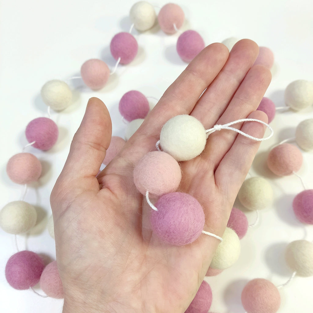 Détail des pompons feutrés de la guirlande guimauve rose dans une main