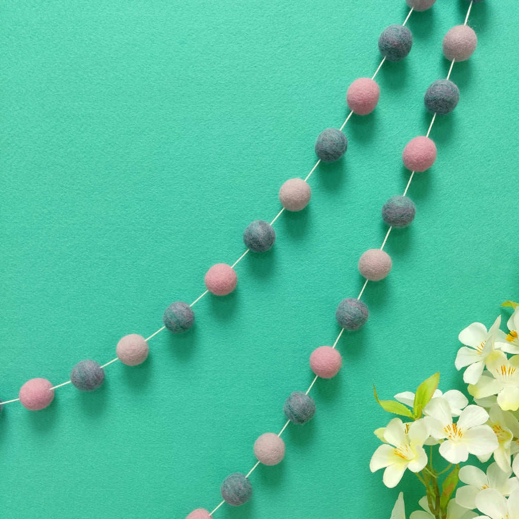 Guirlande de pompons feutrés jardin de lilas suspendue sur un mur, teintes lilas, rose cerisier et mélange turquoise rose lilas