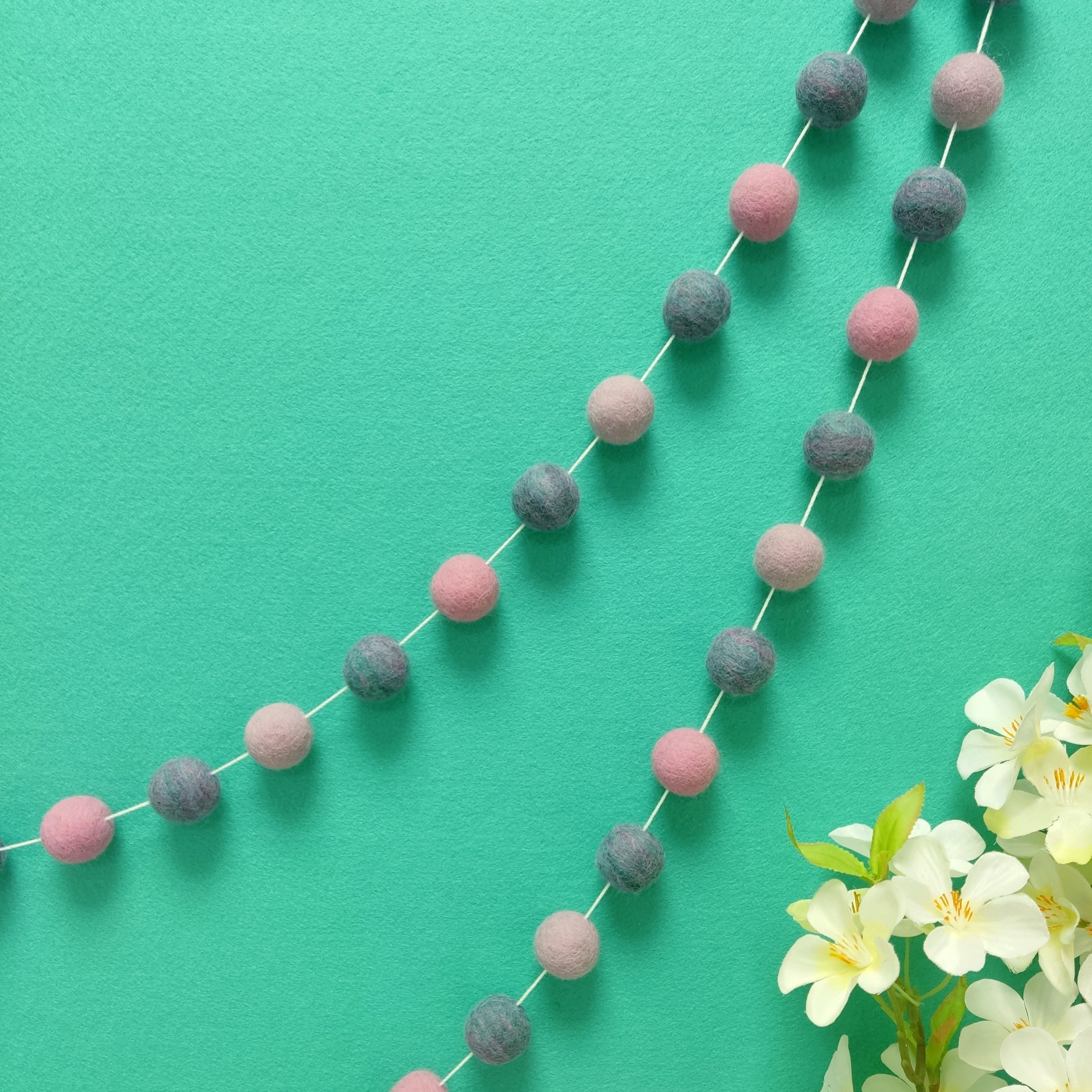 Guirlande de pompons feutrés jardin de lilas suspendue sur un mur, teintes lilas, rose cerisier et mélange turquoise rose lilas