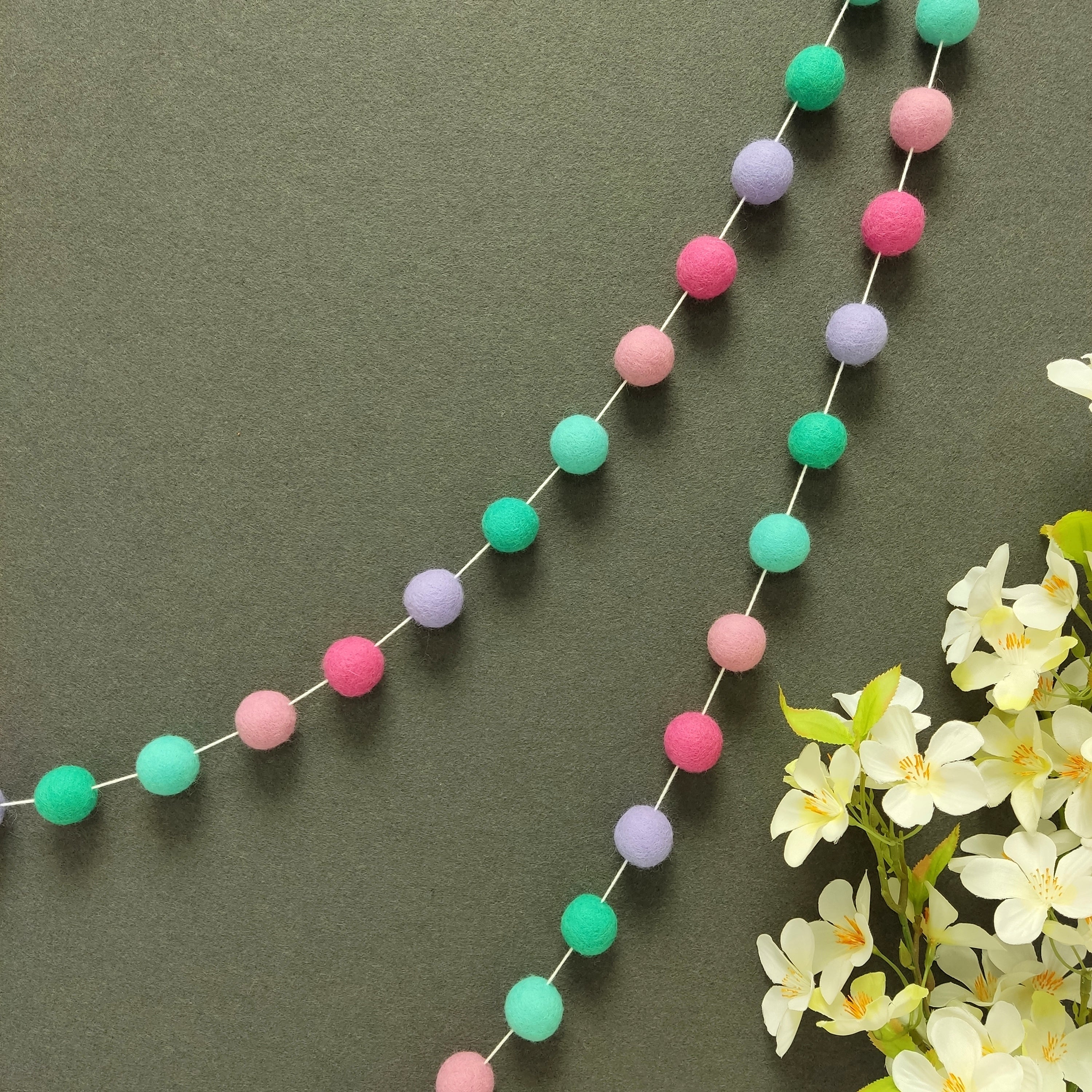 Guirlande de pompons feutrés lagon rose suspendue sur un mur, teintes vert turquoise, vert lagon, lavande, rose bonbon et rose cerisier