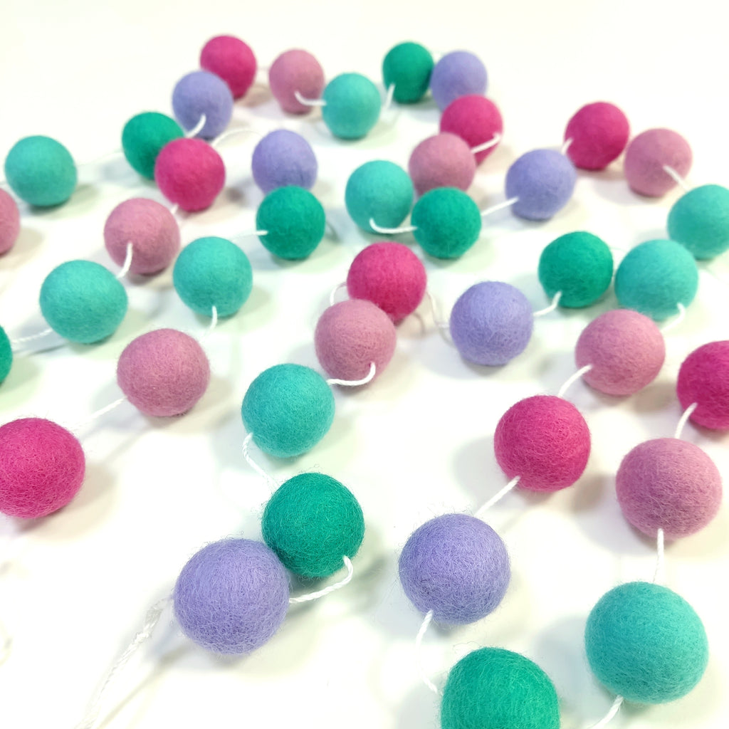 Guirlande de pompons feutrés lagon rose étalée sur une table avec les pompons espacés sur le fil