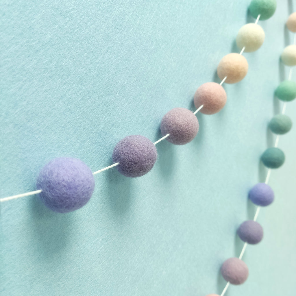 Guirlande de pompons feutrés aux teintes florales lavande, mauve, rose poudré et vert céladon vue de profil