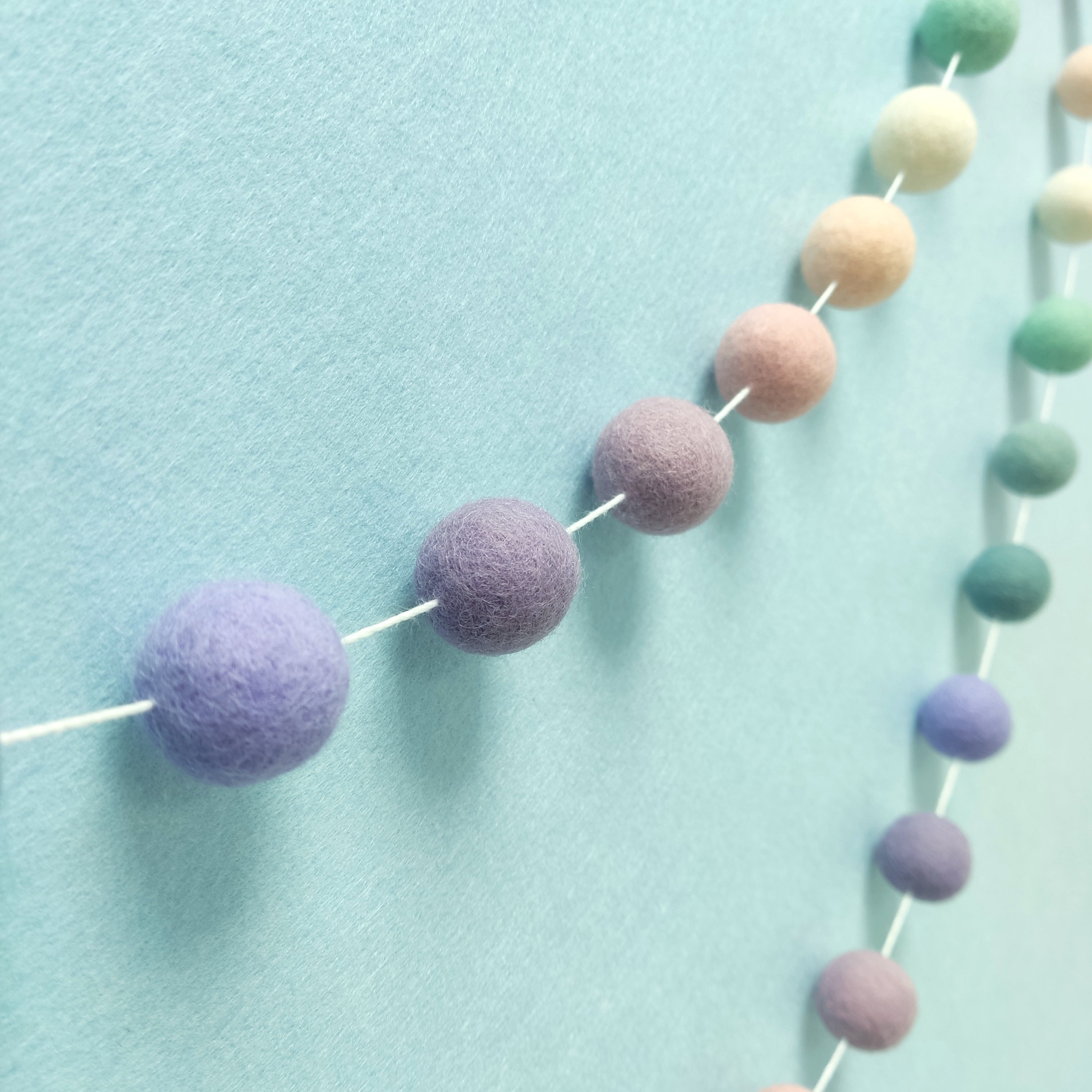 Guirlande de pompons feutrés aux teintes florales lavande, mauve, rose poudré et vert céladon vue de profil
