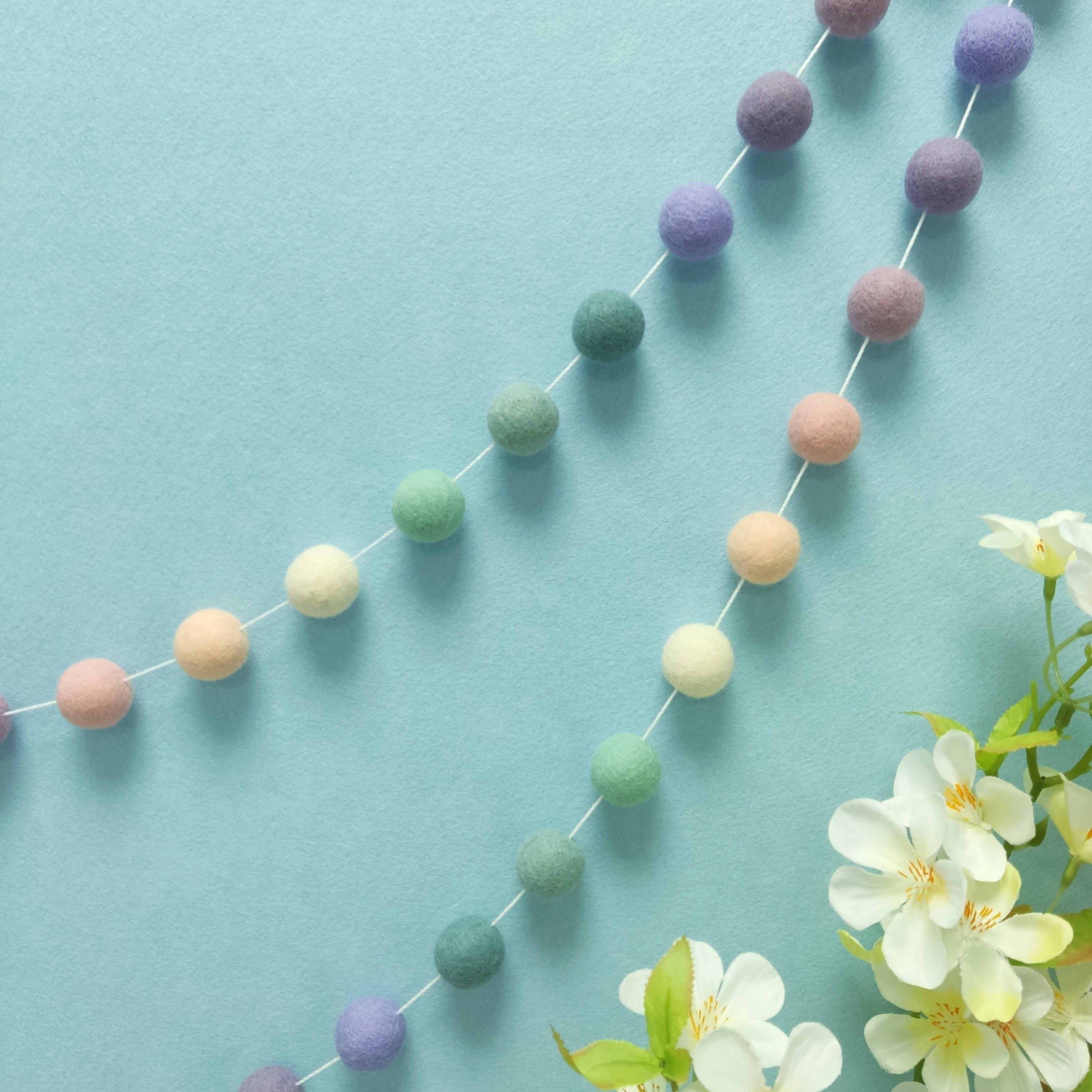 Guirlande de pompons feutrés lavande sauge suspendue sur un mur, teintes blanc crème, rose poudré, lilas, mauve, lavande et vert céladon
