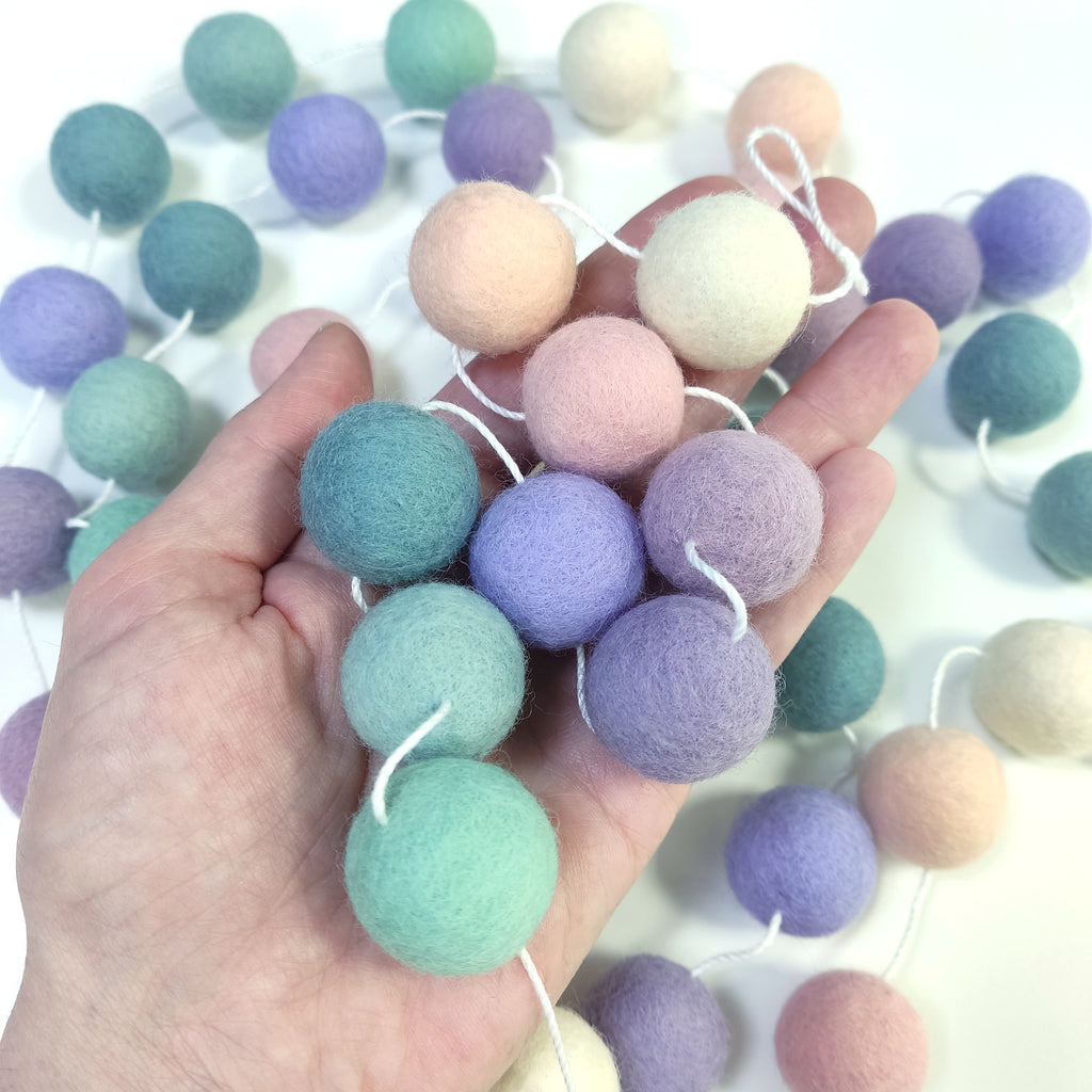 Zoom sur les pompons en laine mérinos lavande sauge dans une main, teintes pastel florales et végétales