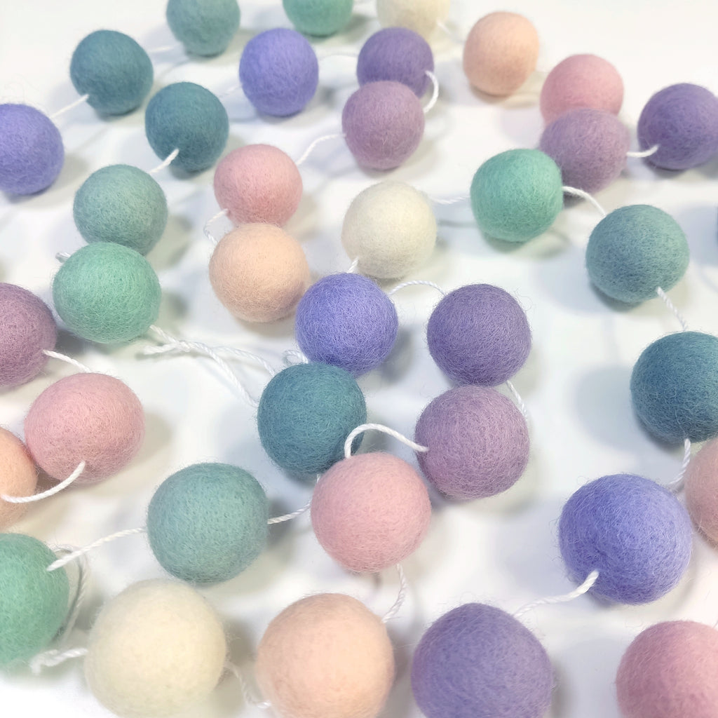 Guirlande de pompons feutrés lavande sauge étalée sur une table avec les pompons espacés sur le fil