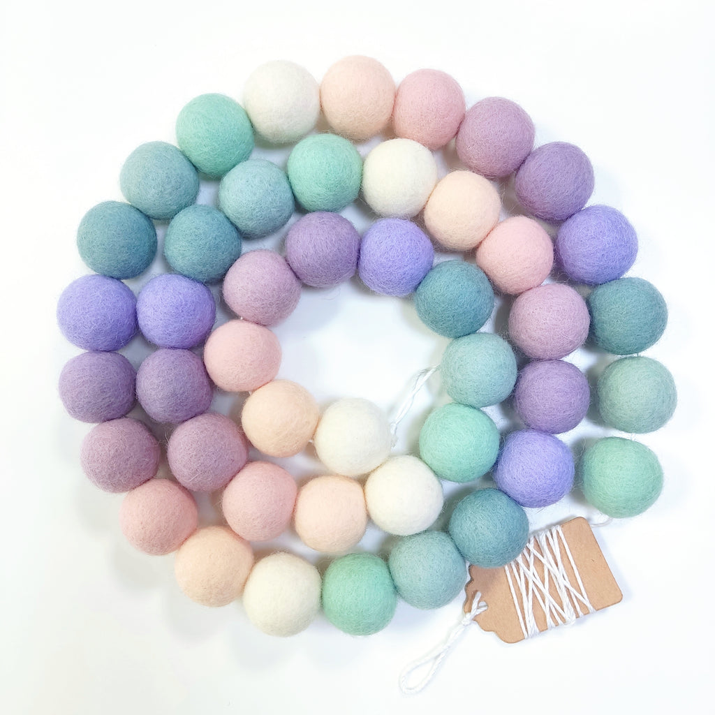 Guirlande de pompons feutrés lavande sauge disposée en spirale sur fond blanc