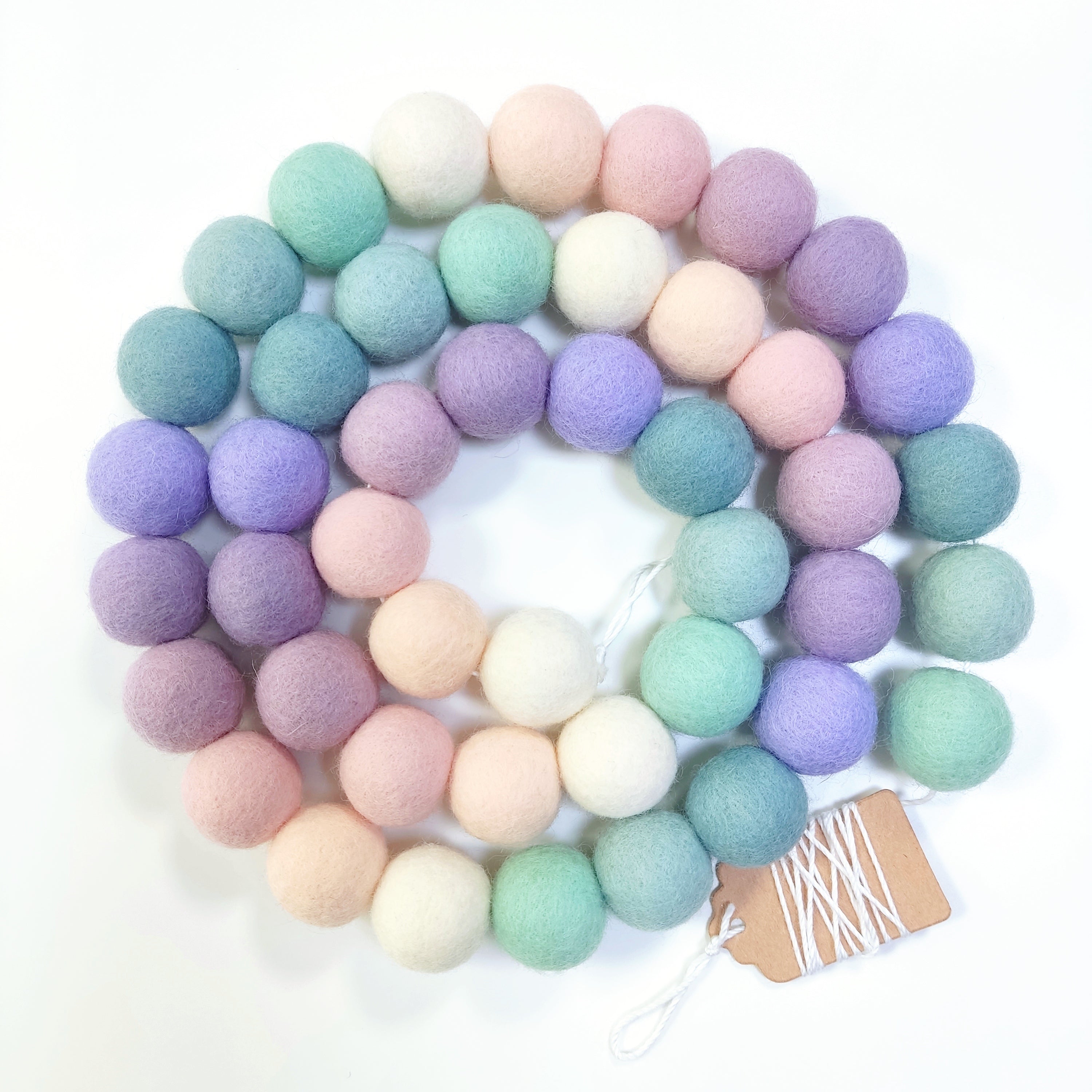 Guirlande de pompons feutrés lavande sauge disposée en spirale sur fond blanc