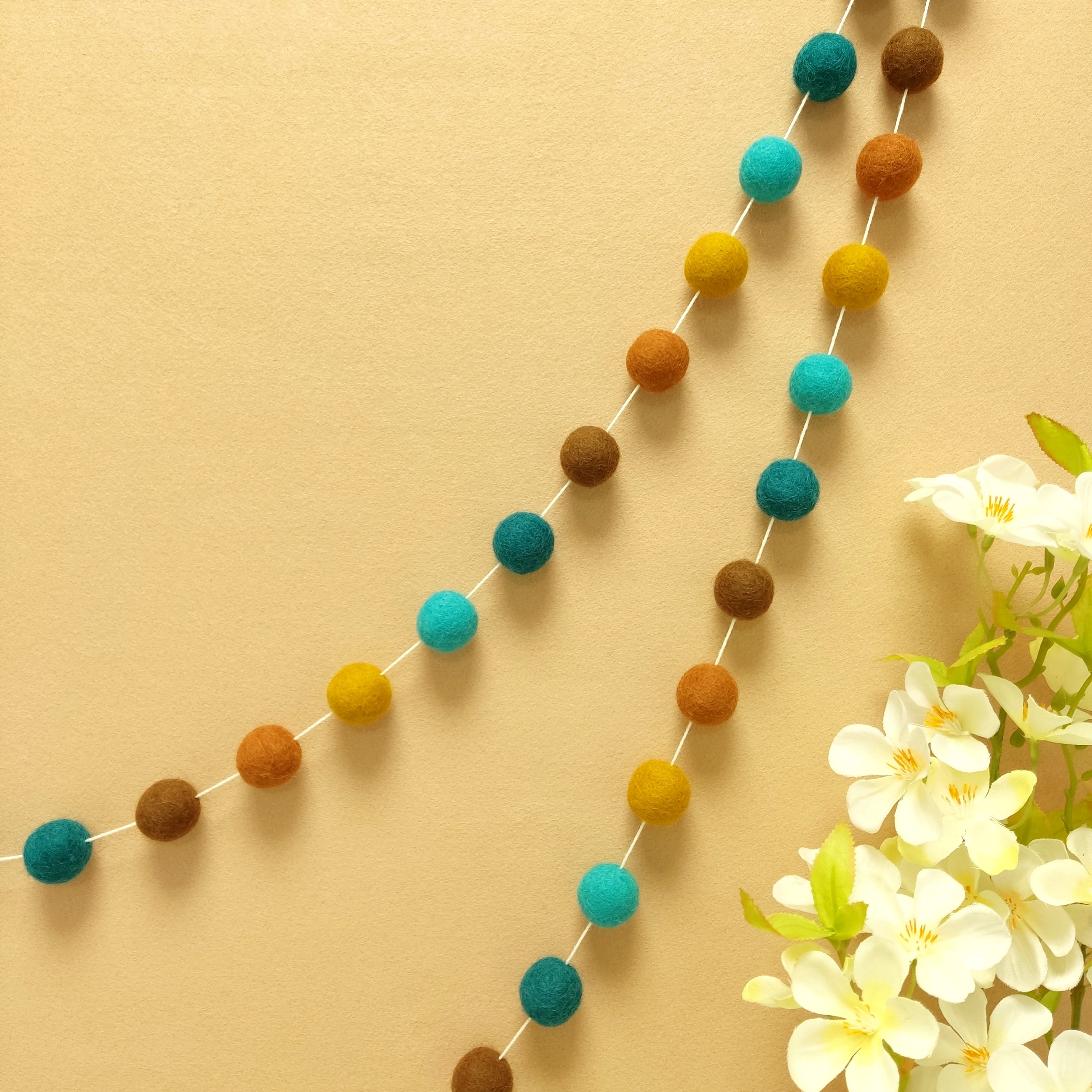 Guirlande de pompons feutrés Marrakech suspendue sur un mur, teintes brun noisette, terracotta cuivré, ocre doré, bleu turquoise et vert canard