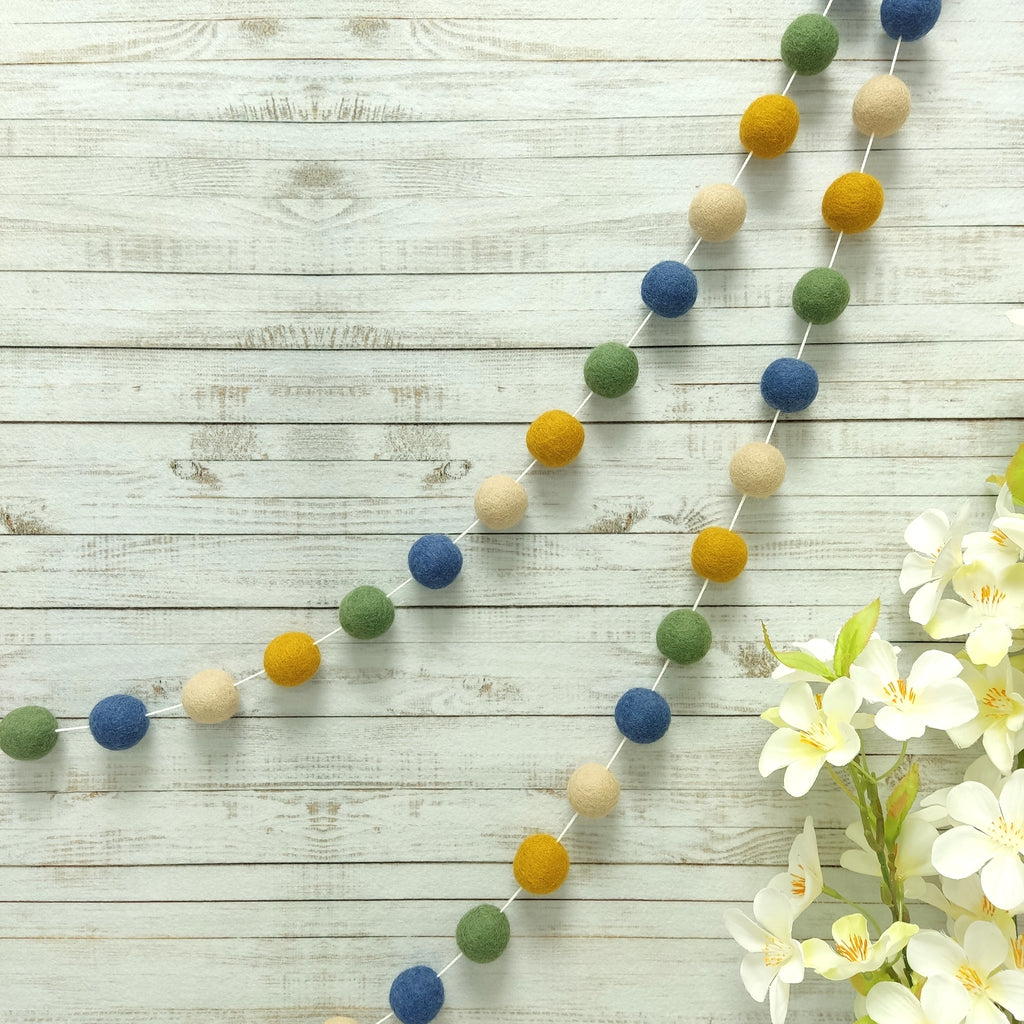Guirlande de pompons matcha, bleu beige et ocre suspendue sur un mur