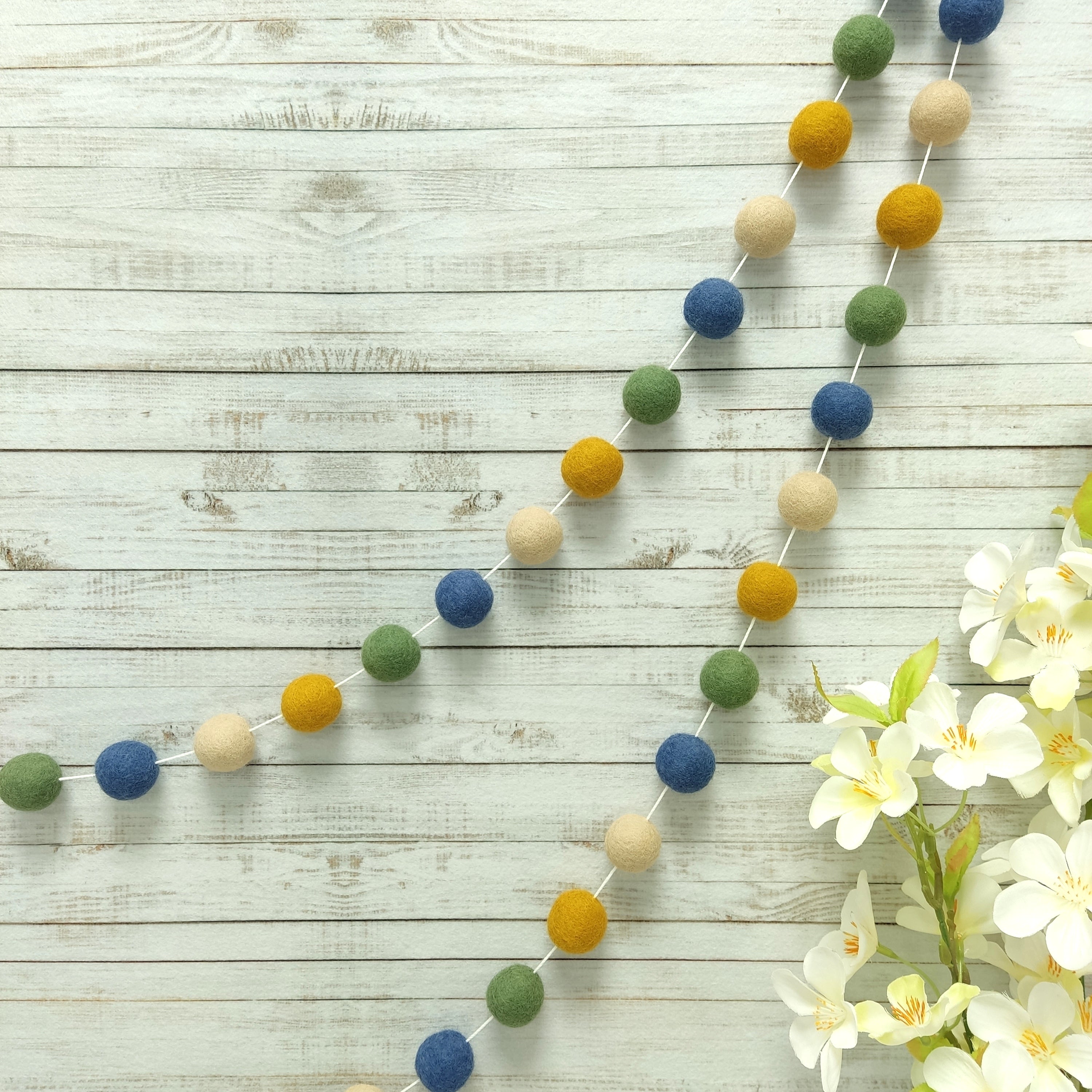 Guirlande de pompons matcha, bleu beige et ocre suspendue sur un mur