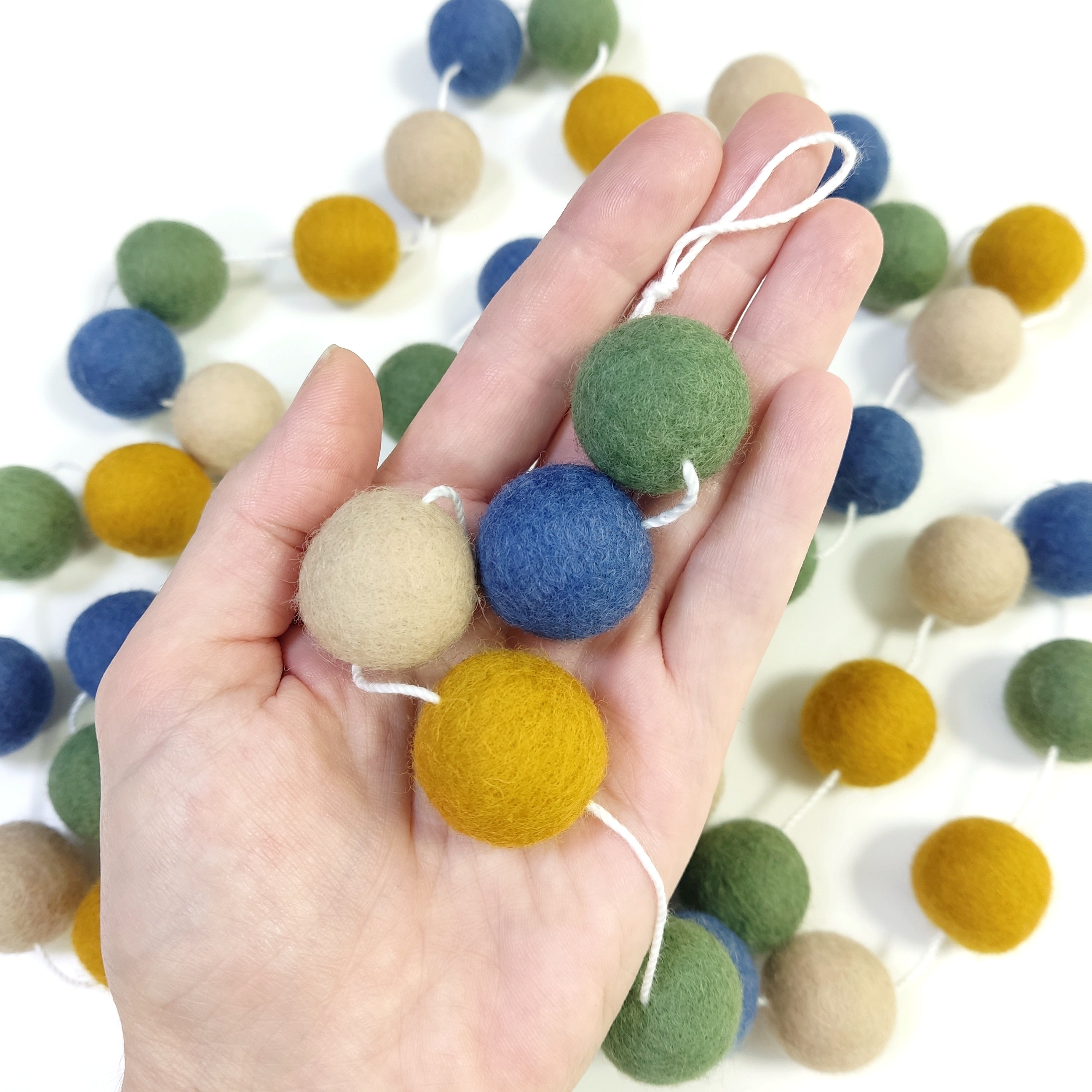Détail des pompons en laine feutrée ocre doré, bleu denim, beige poudré et vert matcha dans une main