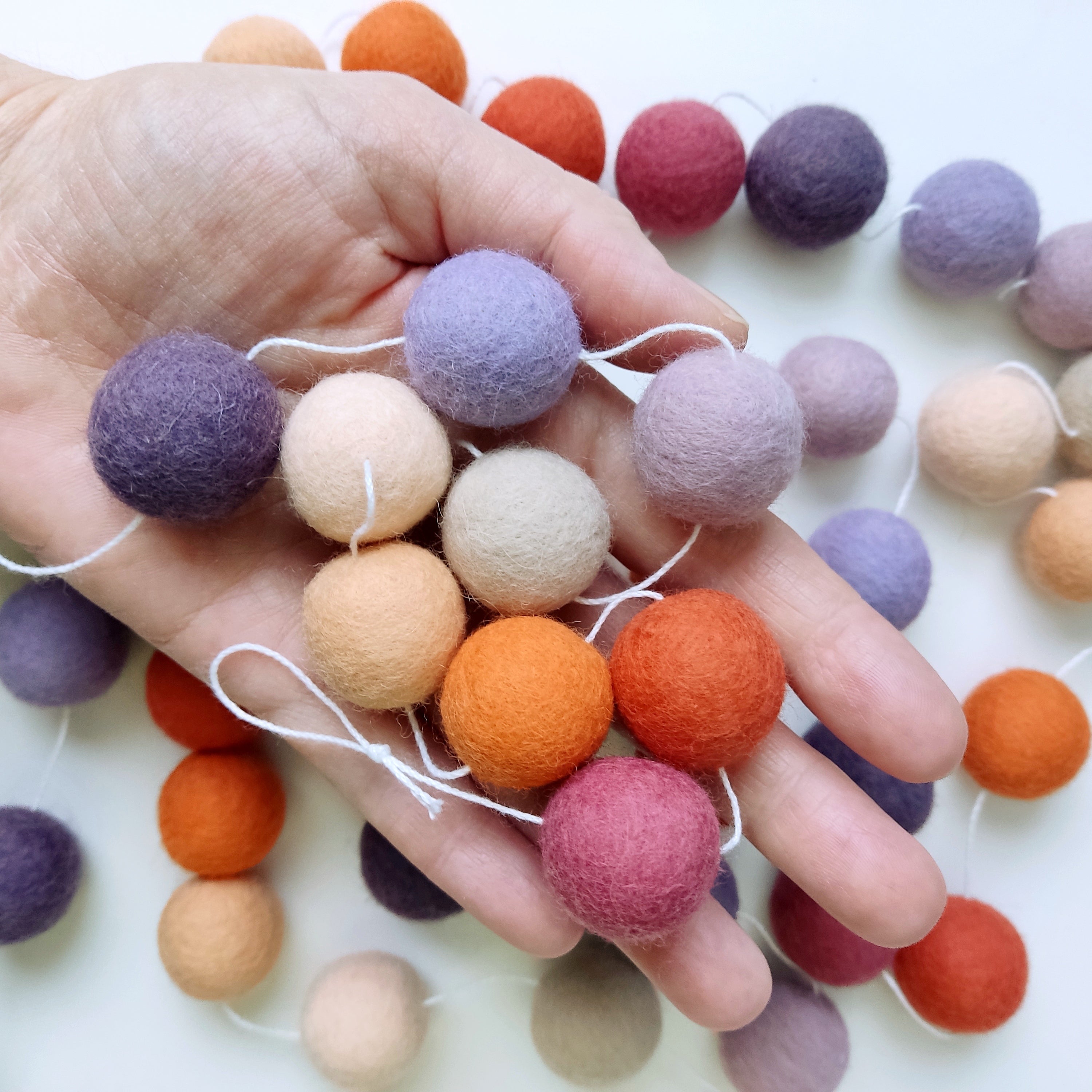 Zoom sur les pompons en feutre de laine mérinos de couleur mauve, rose, violet, orange et beige dans une main