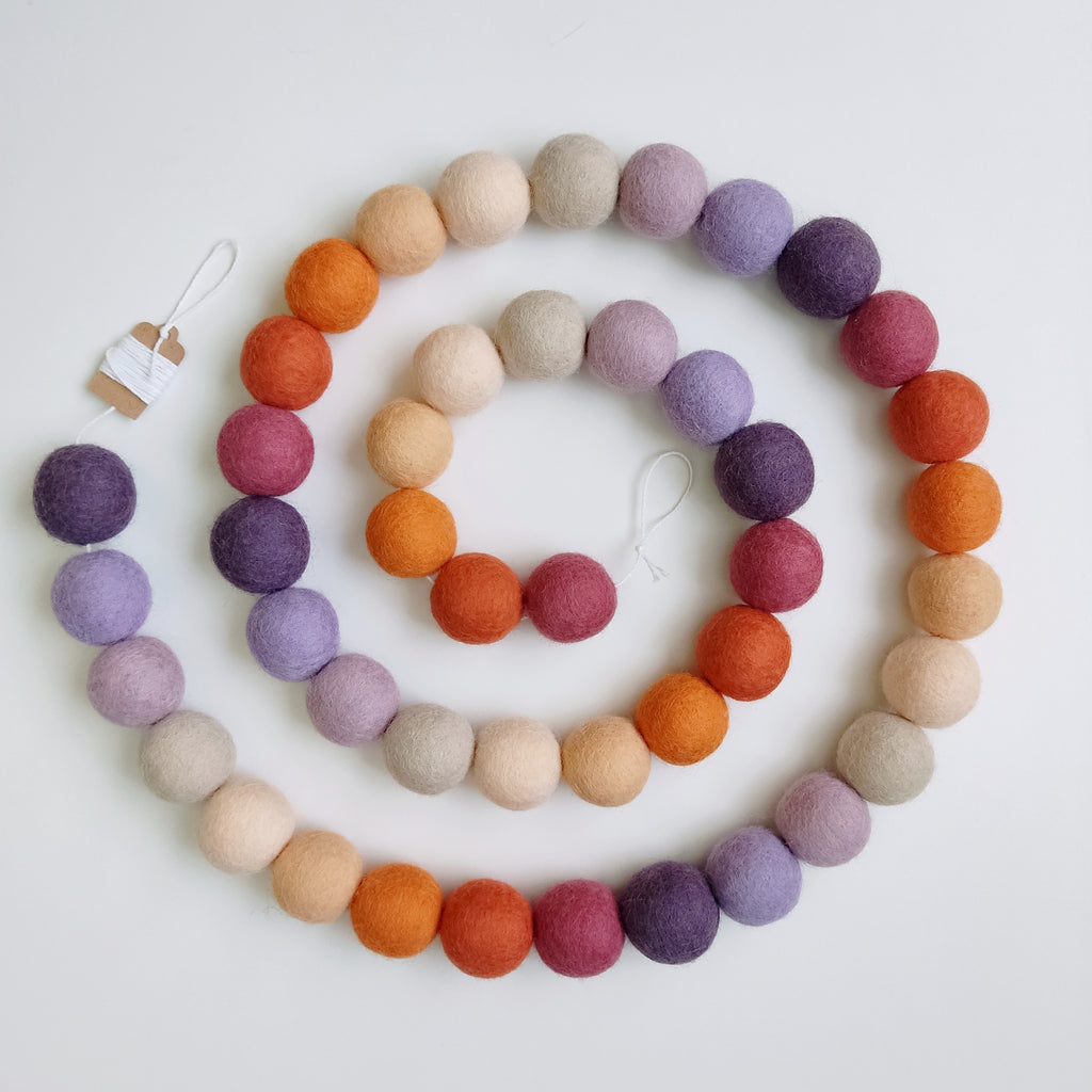 Guirlande en boules de feutre beige, mauve, orange, violet et rose posée en spirale sur une table