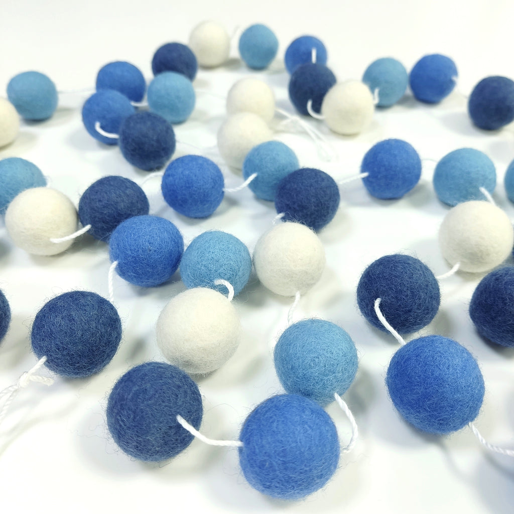 Guirlande de boule de laine feutrée couleurs bleu et blanc étalée sur une table avec les pompons espacés