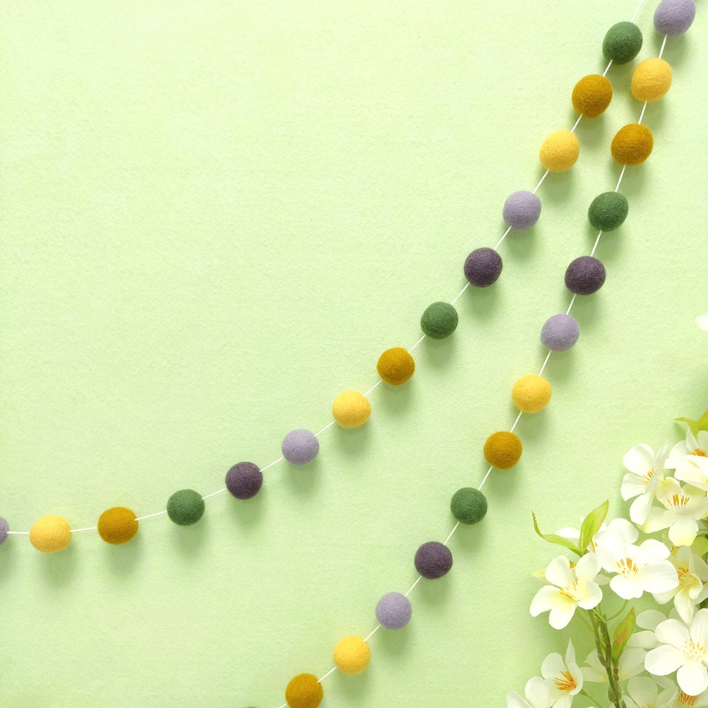 Guirlande de pompons feutrés miel lavande suspendue sur un mur, teintes jaune maïs, jaune ocre, vert matcha, mauve et violet améthyste
