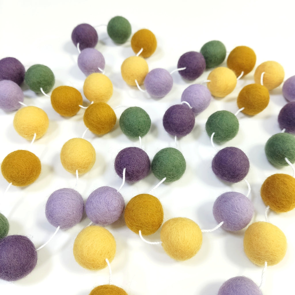 Guirlande de pompons feutrés miel lavande étalée sur une table avec les pompons espacés sur le fil