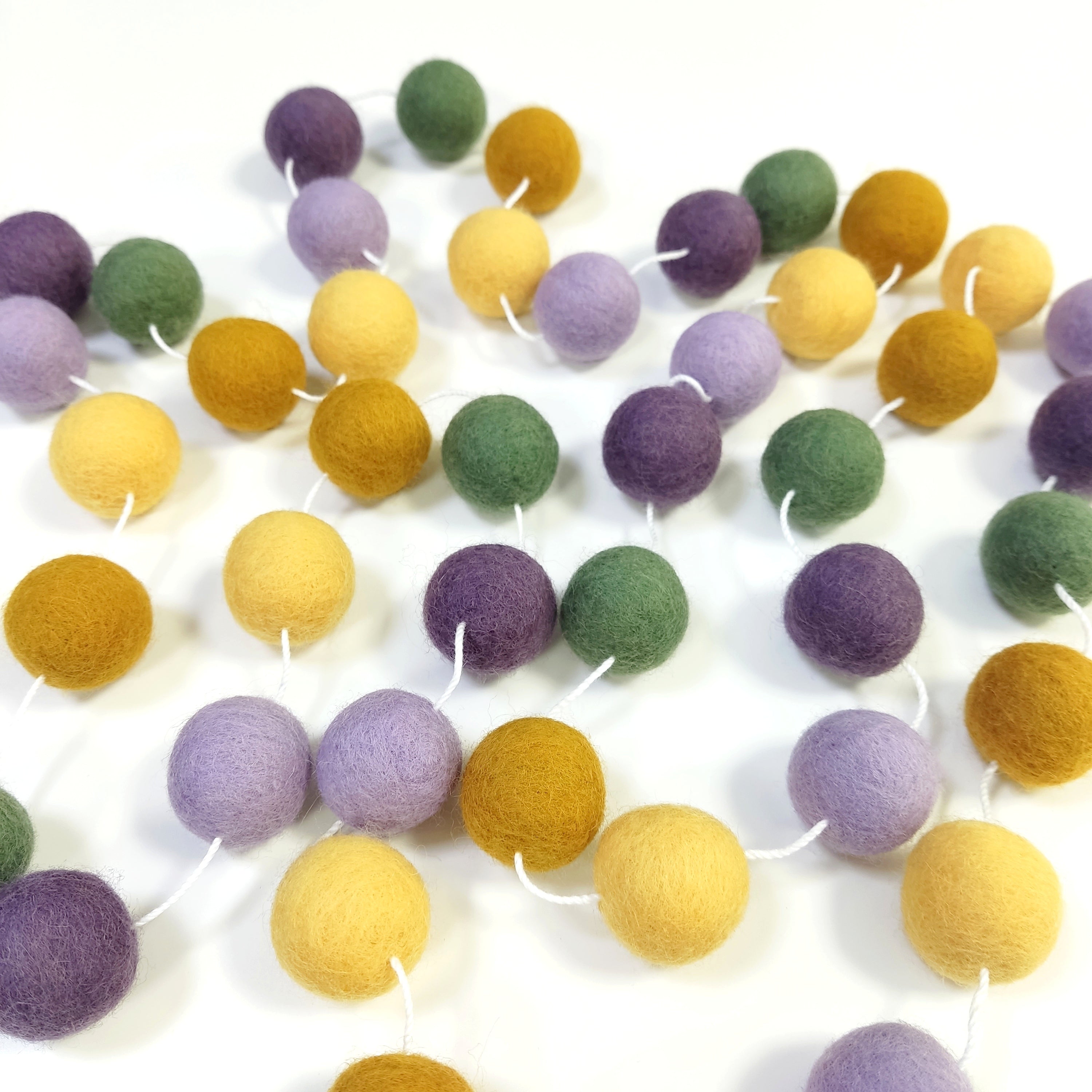 Guirlande de pompons feutrés miel lavande étalée sur une table avec les pompons espacés sur le fil