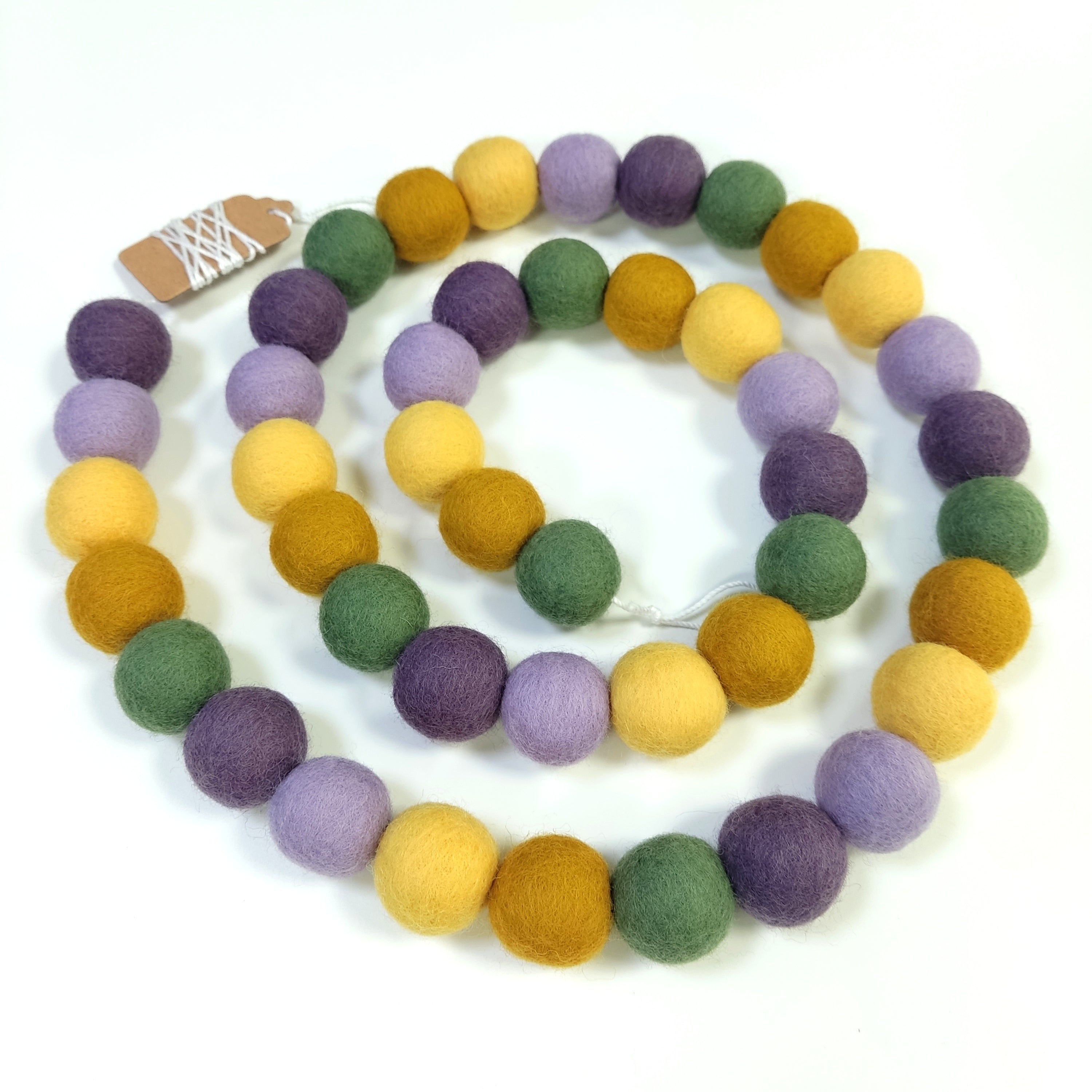 Guirlande de pompons feutrés miel lavande disposée en spirale sur fond blanc