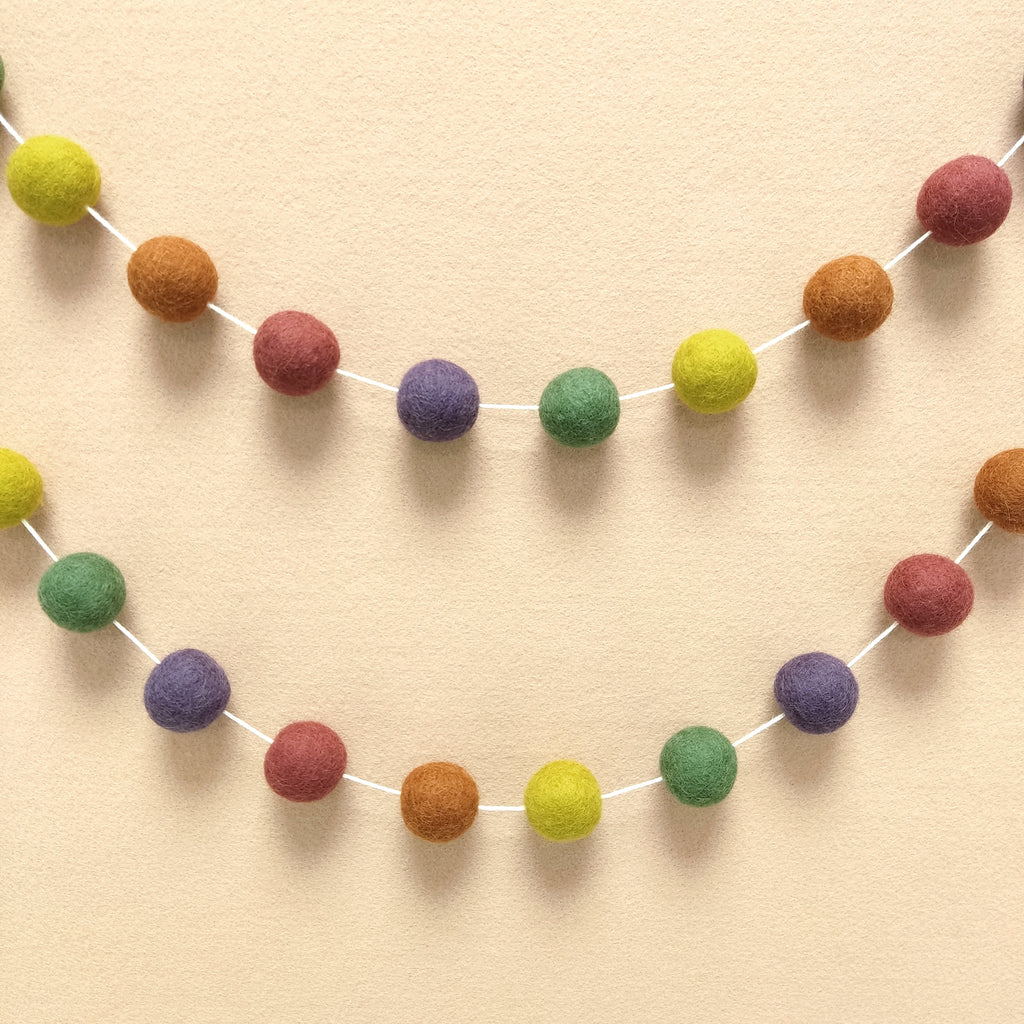 Guirlande en boules de feutre vert rose terracotta violet jaune accrochée en arc de cercle sur un mur