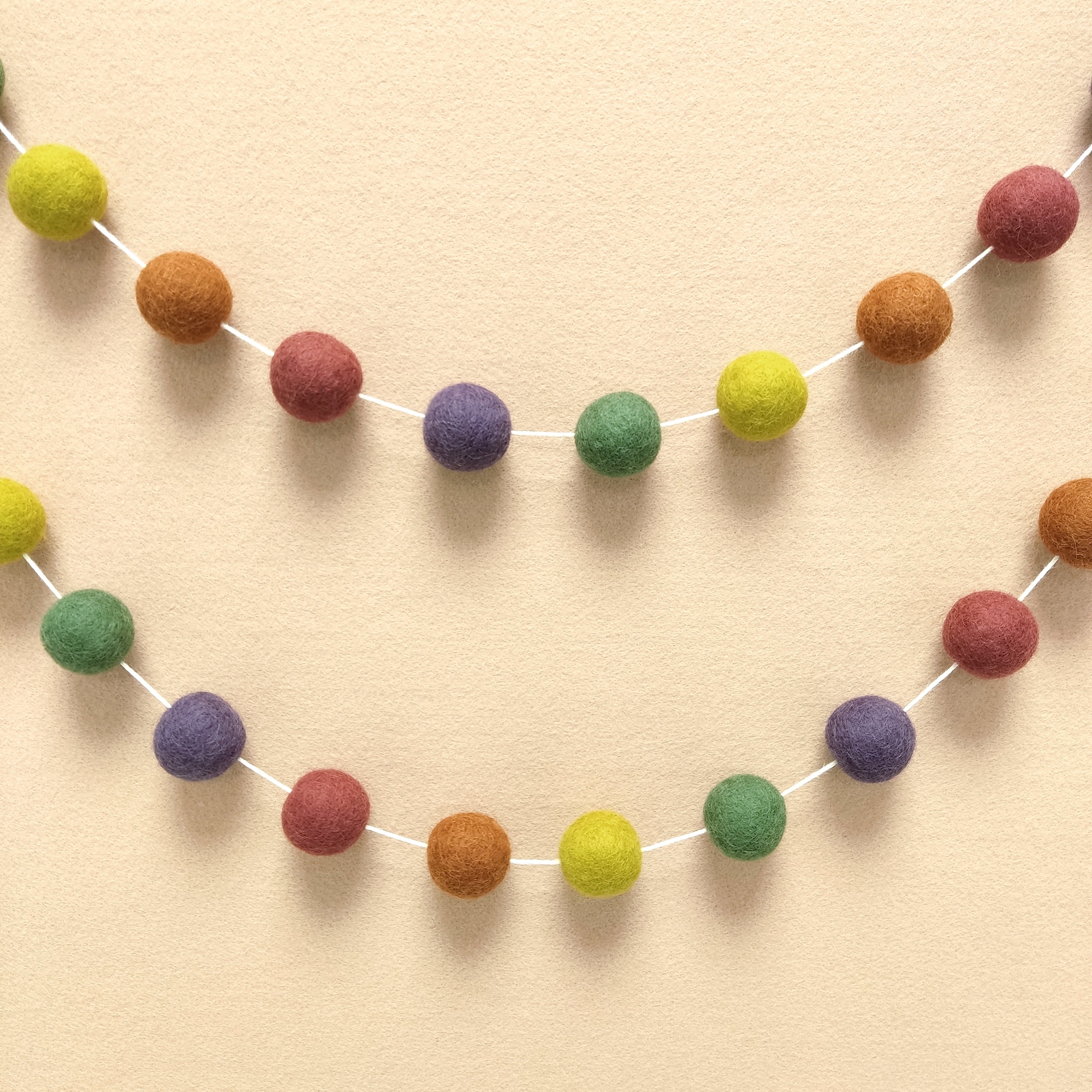Guirlande en boules de feutre vert rose terracotta violet jaune accrochée en arc de cercle sur un mur
