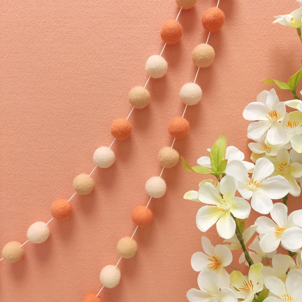 Guirlande de pompons feutrés orange flamingo, pêche et beige rosé suspendue sur un mur, faite main en France