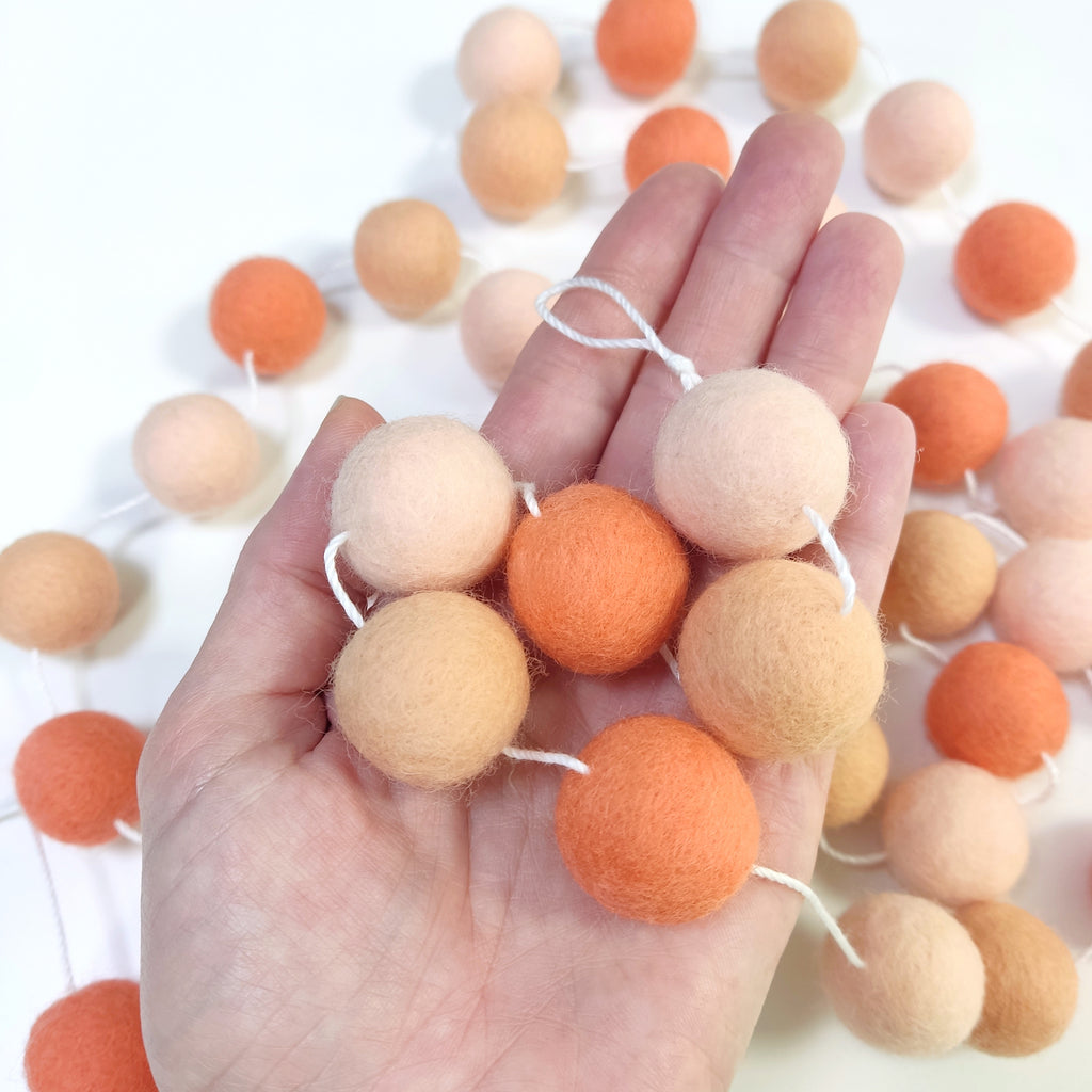 Zoom sur les pompons beige rosé, orange pêche, orange flamingo de la guirlande dans une main