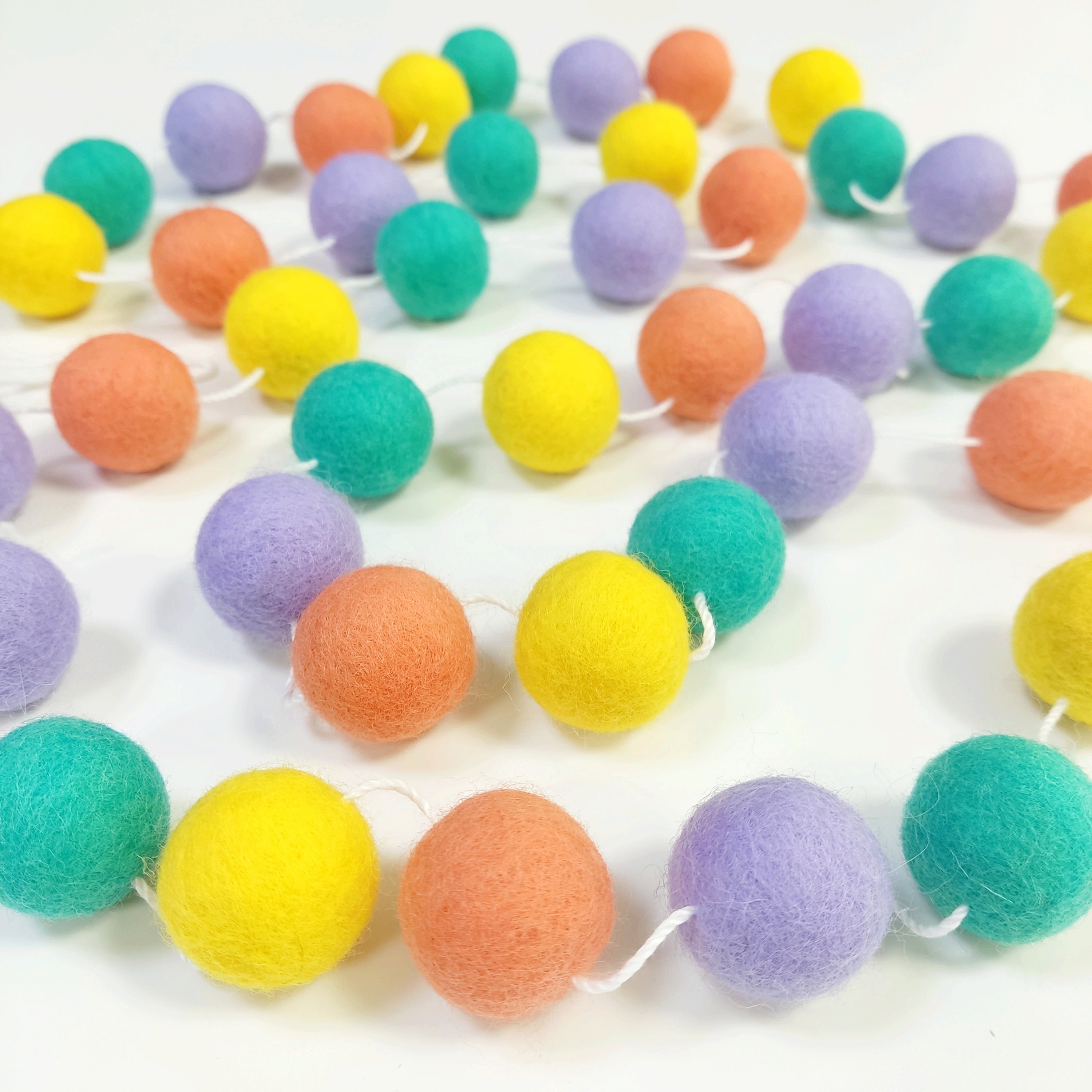 Guirlande de boules de laine feutrée pastel pop posée sur une table avec les pompons espacés