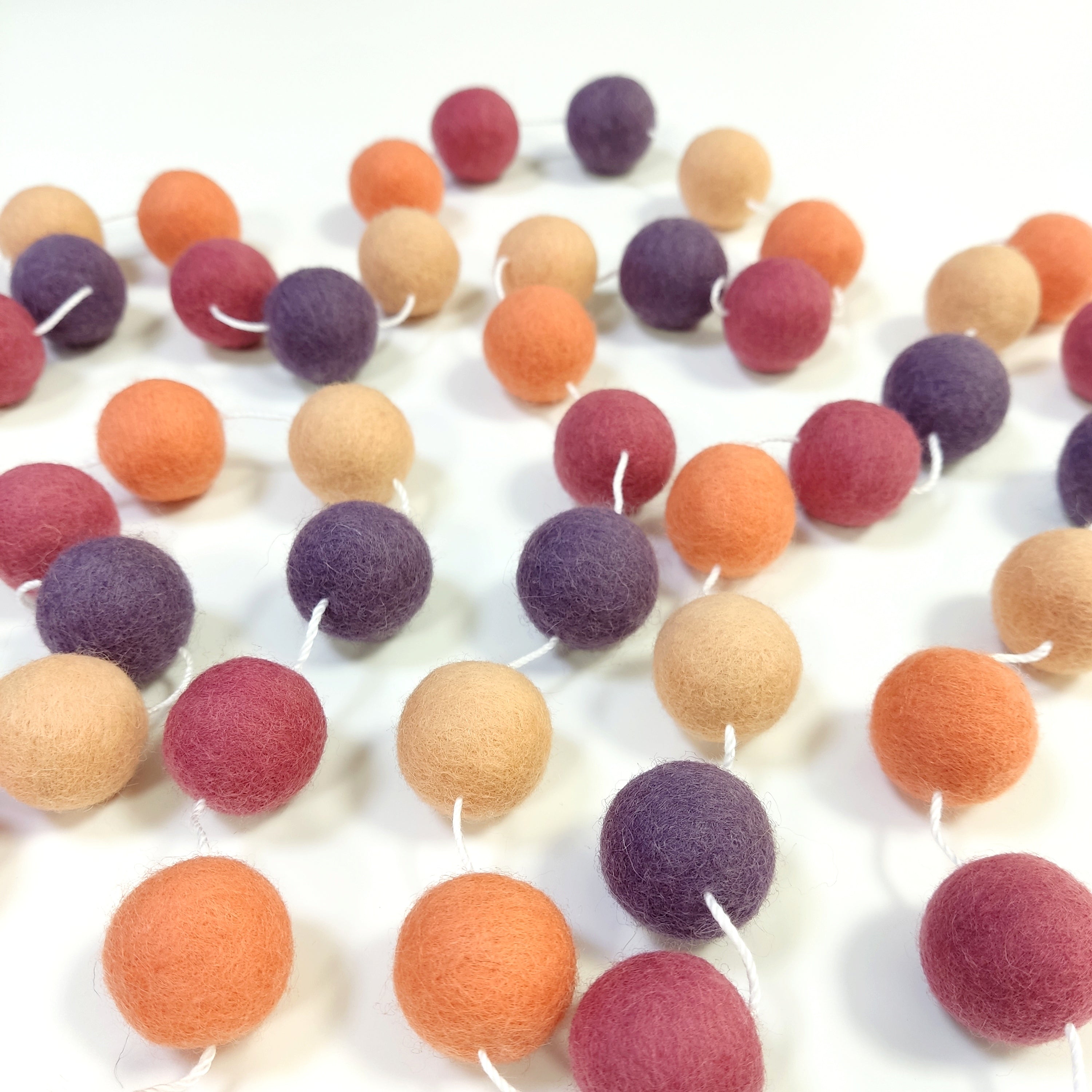 Guirlande de pompons feutrés pêche améthyste étalée sur une table avec les pompons espacés sur le fil
