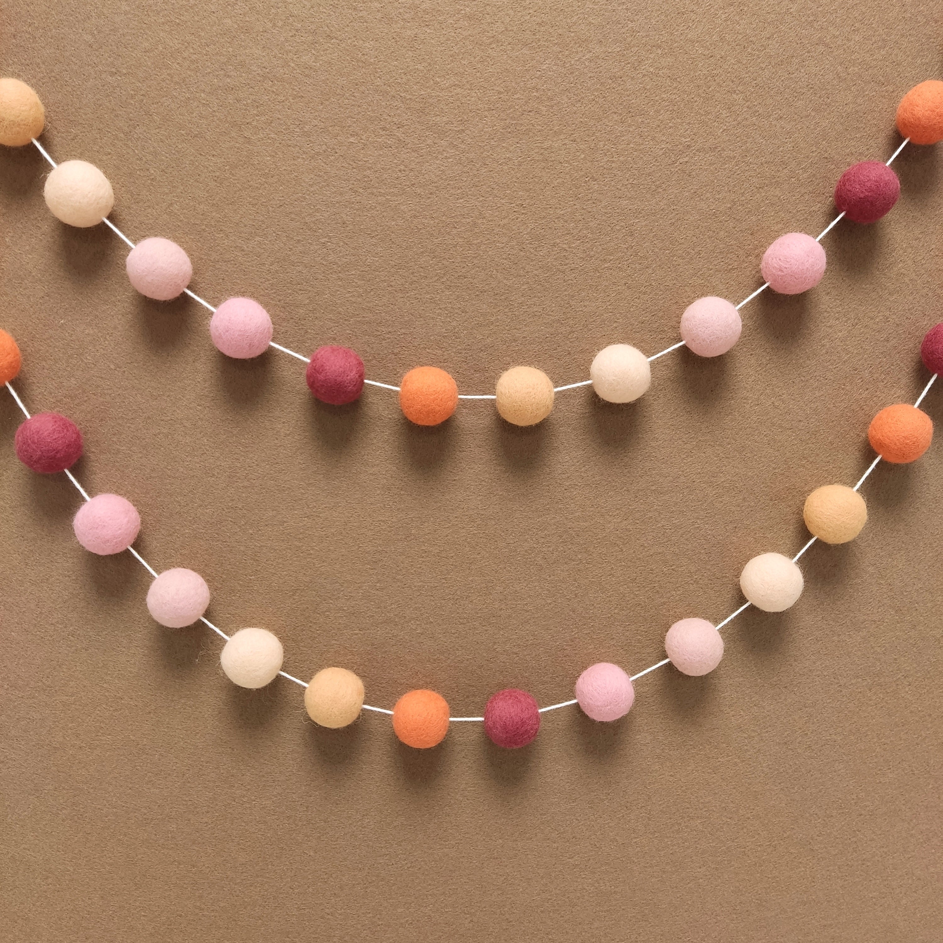 Double guirlande en boules de feutre orange pêche rose clair et rose fixées en arc de cercle sur un mur de chambre