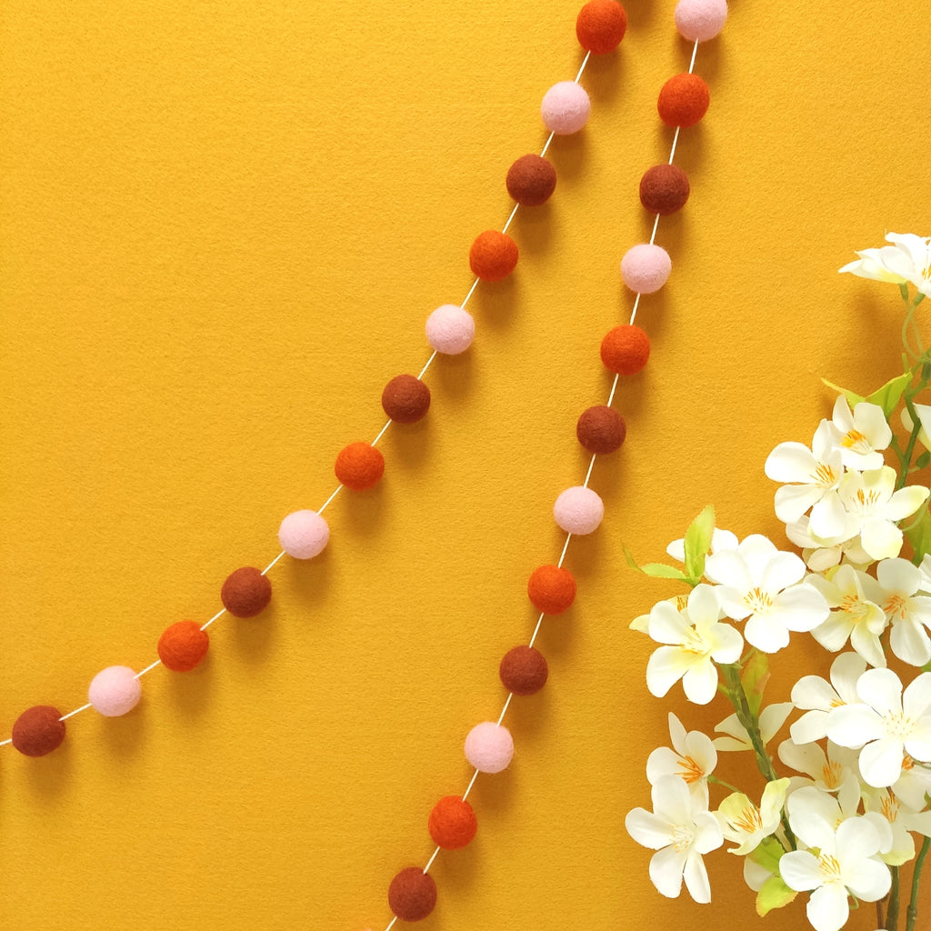 Guirlande de pompons feutrés pétale terracotta suspendue sur un mur, teintes terracotta rouge, orange mandarine et rose poudré