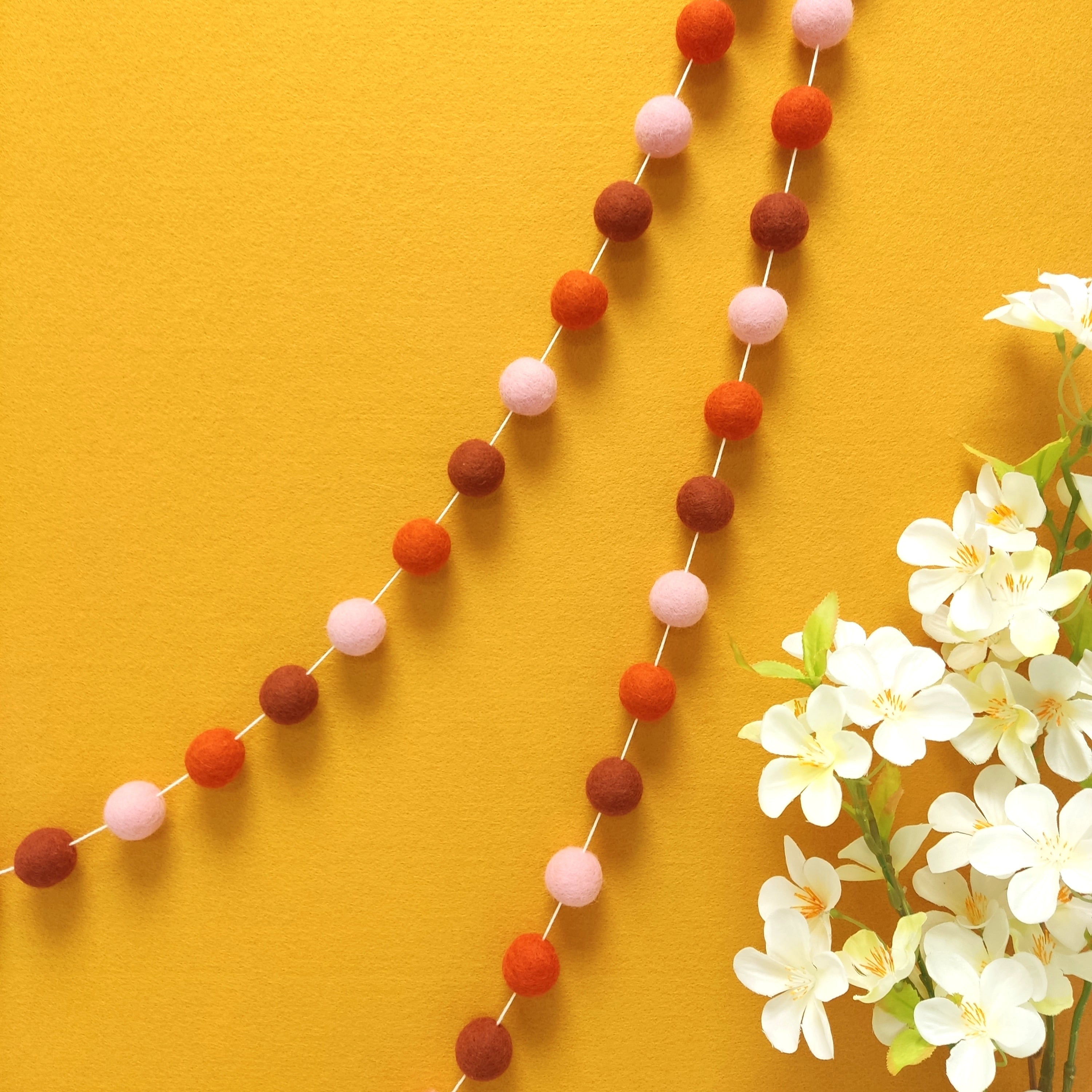 Guirlande de pompons feutrés pétale terracotta suspendue sur un mur, teintes terracotta rouge, orange mandarine et rose poudré