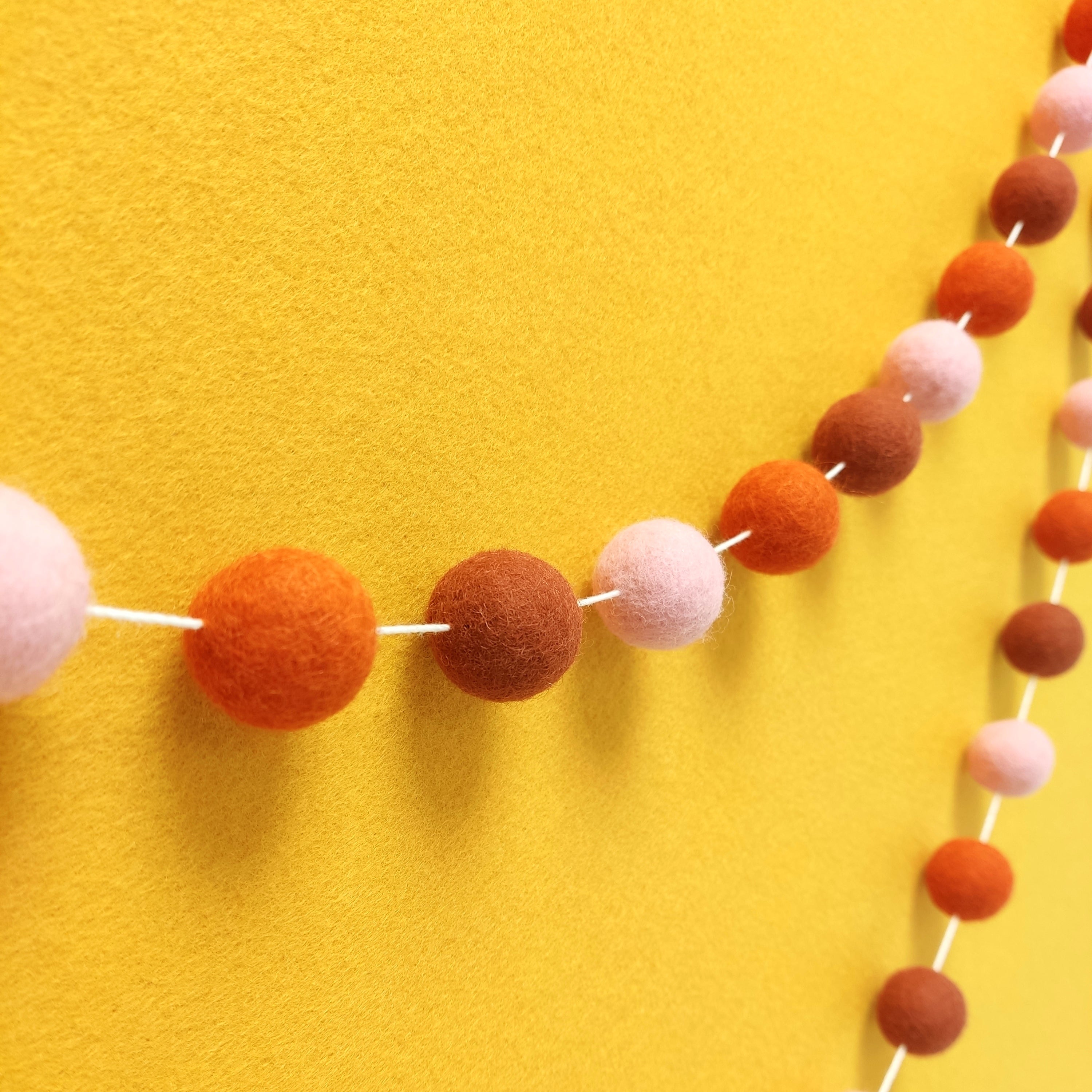 Guirlande de pompons feutrés aux teintes automnales terracotta rouge, orange mandarine et rose poudré vue de profil