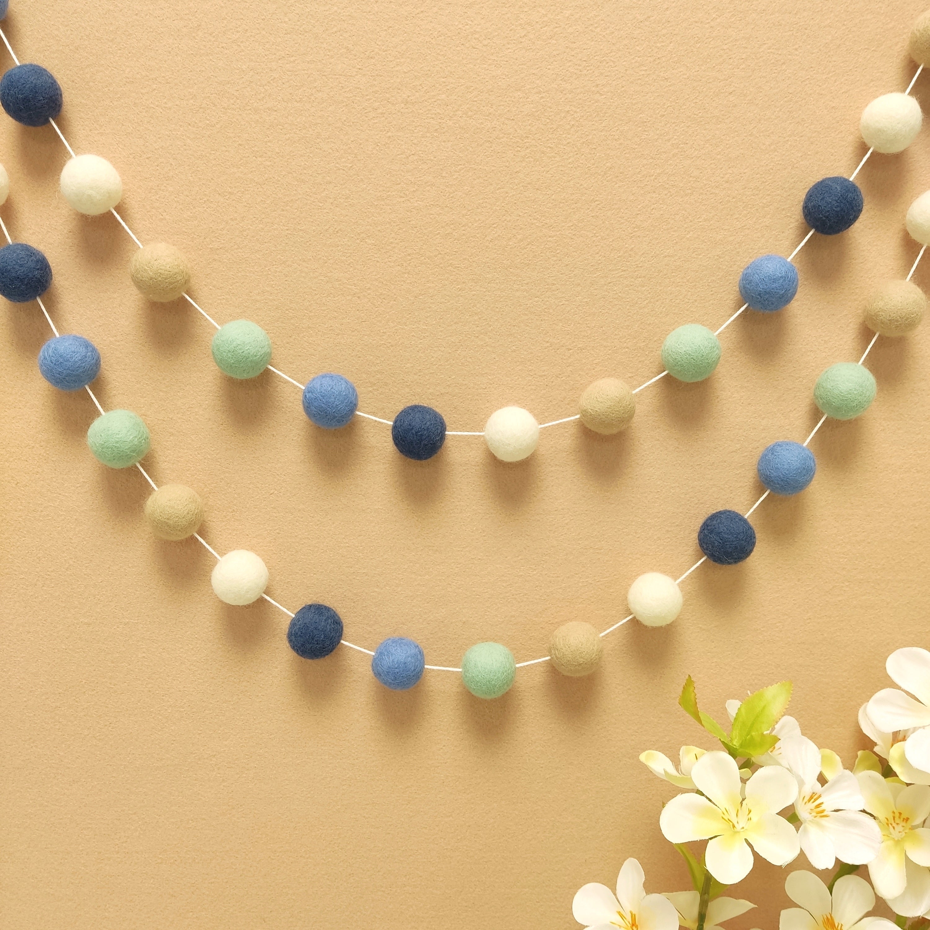 Guirlande de pompons feutrés riviera aux teintes bleu denim, ciel beige blanc et vert menthe accrochée sur un mur