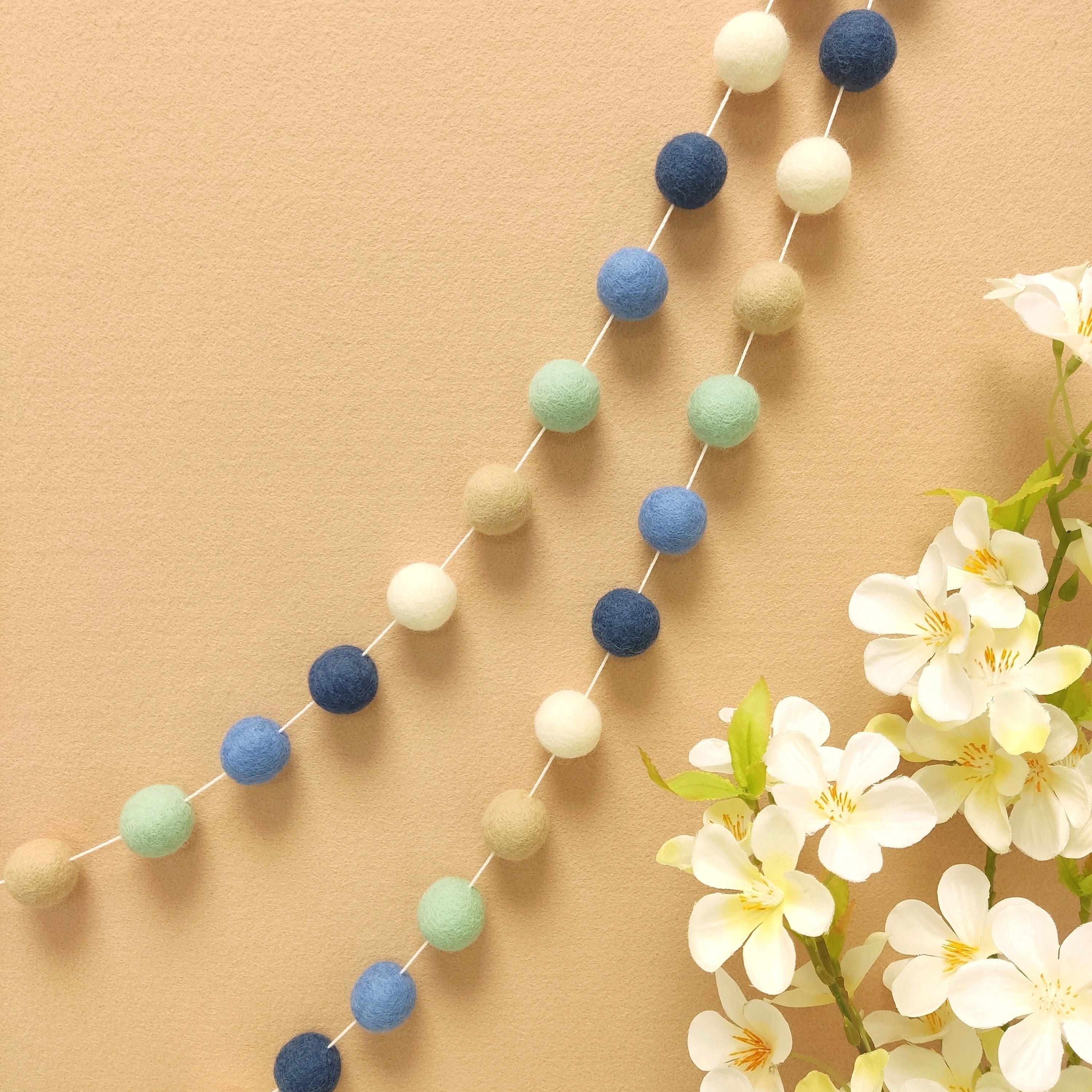 Guirlande de pompons en feutre de laine mérinos beige vert bleu blanc suspendue sur un mur