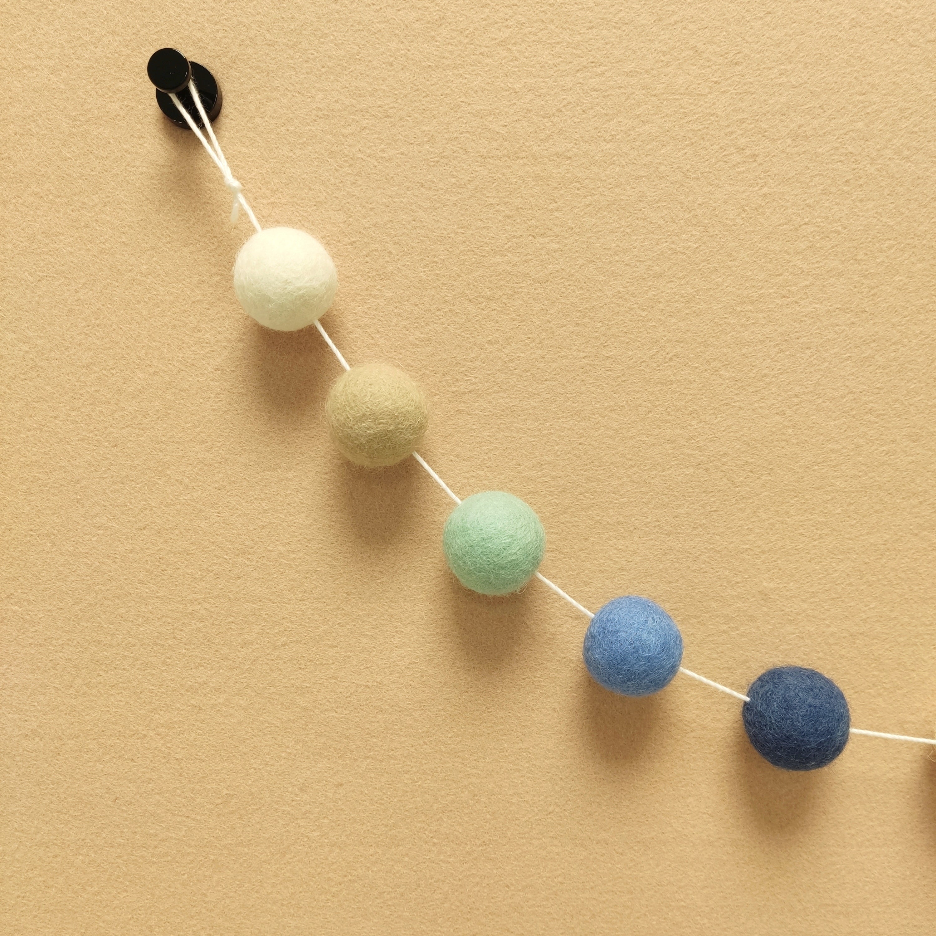 Zoom sur les pompons feutrés blanc, beige poudré, vert menthe, bleu chambray et bleu denim de la guirlande riviera avec la boucle de fixation